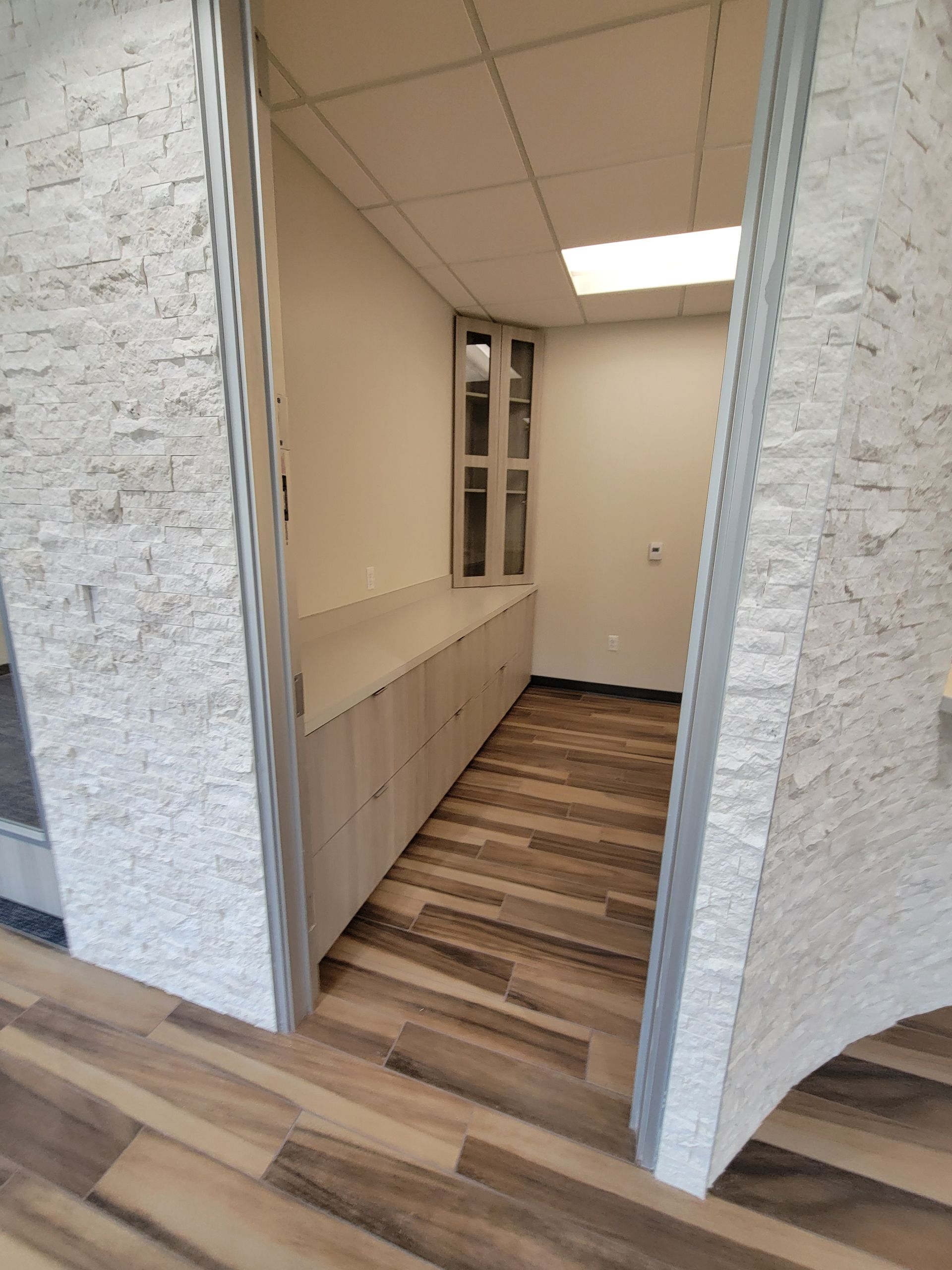 View into a room with a counter, cabinets, window, and wood-look flooring; light-colored stone wall accents.