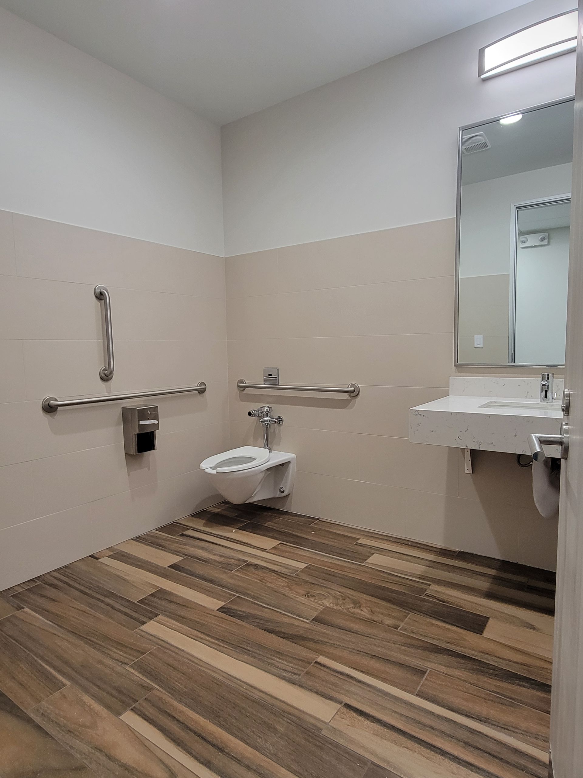 Accessible restroom with wood-look floor, wall-mounted toilet, sink, and grab bars.