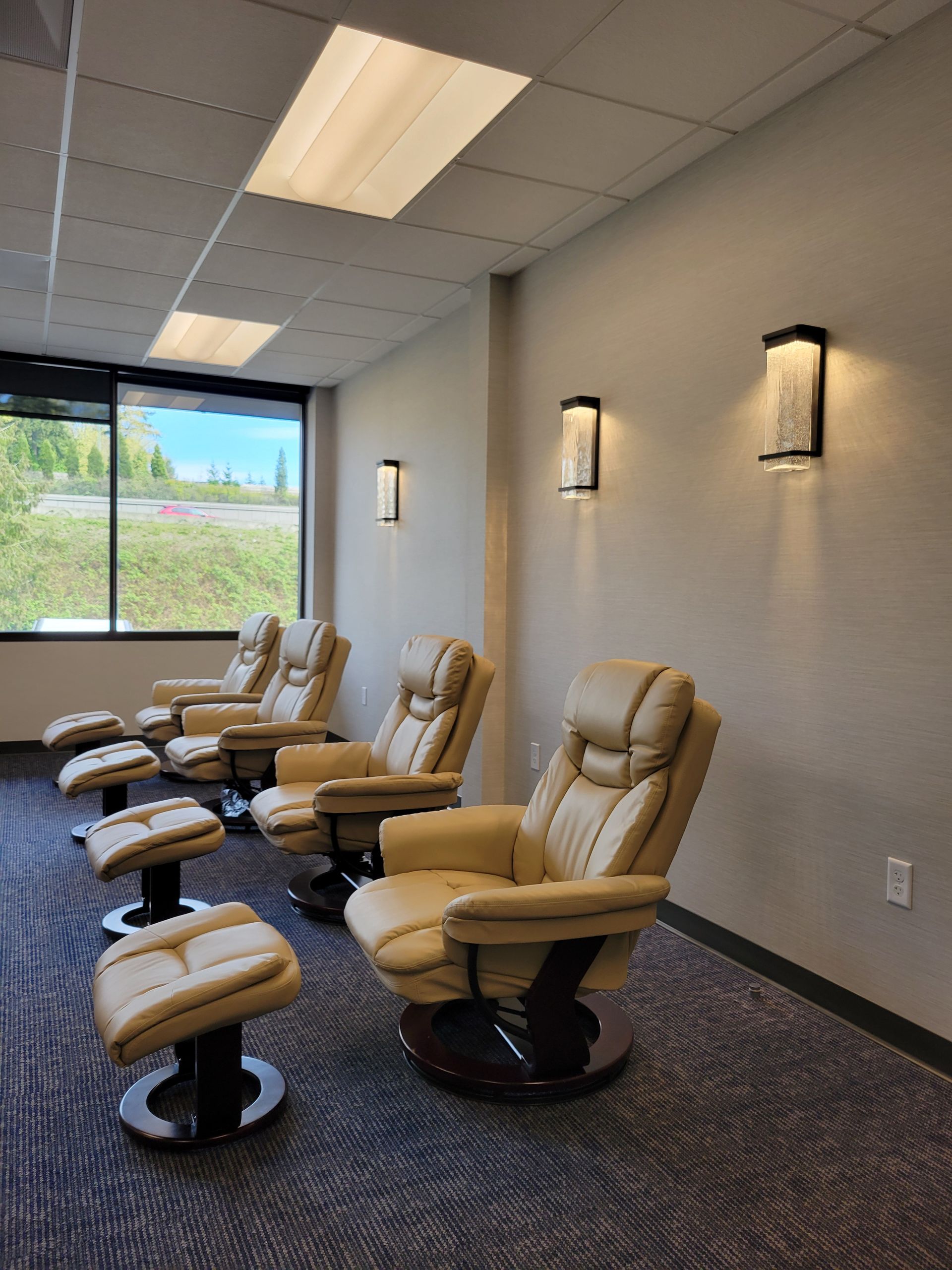 Three beige reclining armchairs with matching footstools arranged in a row in a modern, carpeted room with wall lighting.