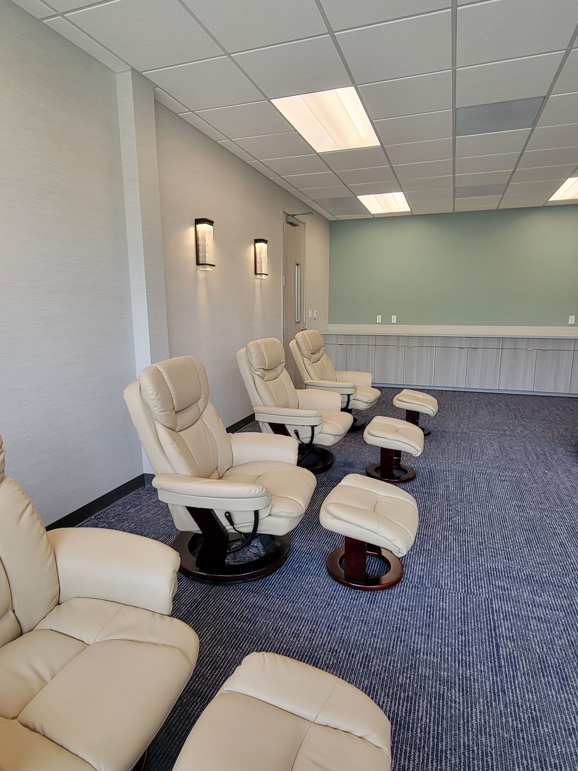 Lounge area with cream-colored recliners and footrests on patterned blue carpet, against light walls and a teal accent wall.