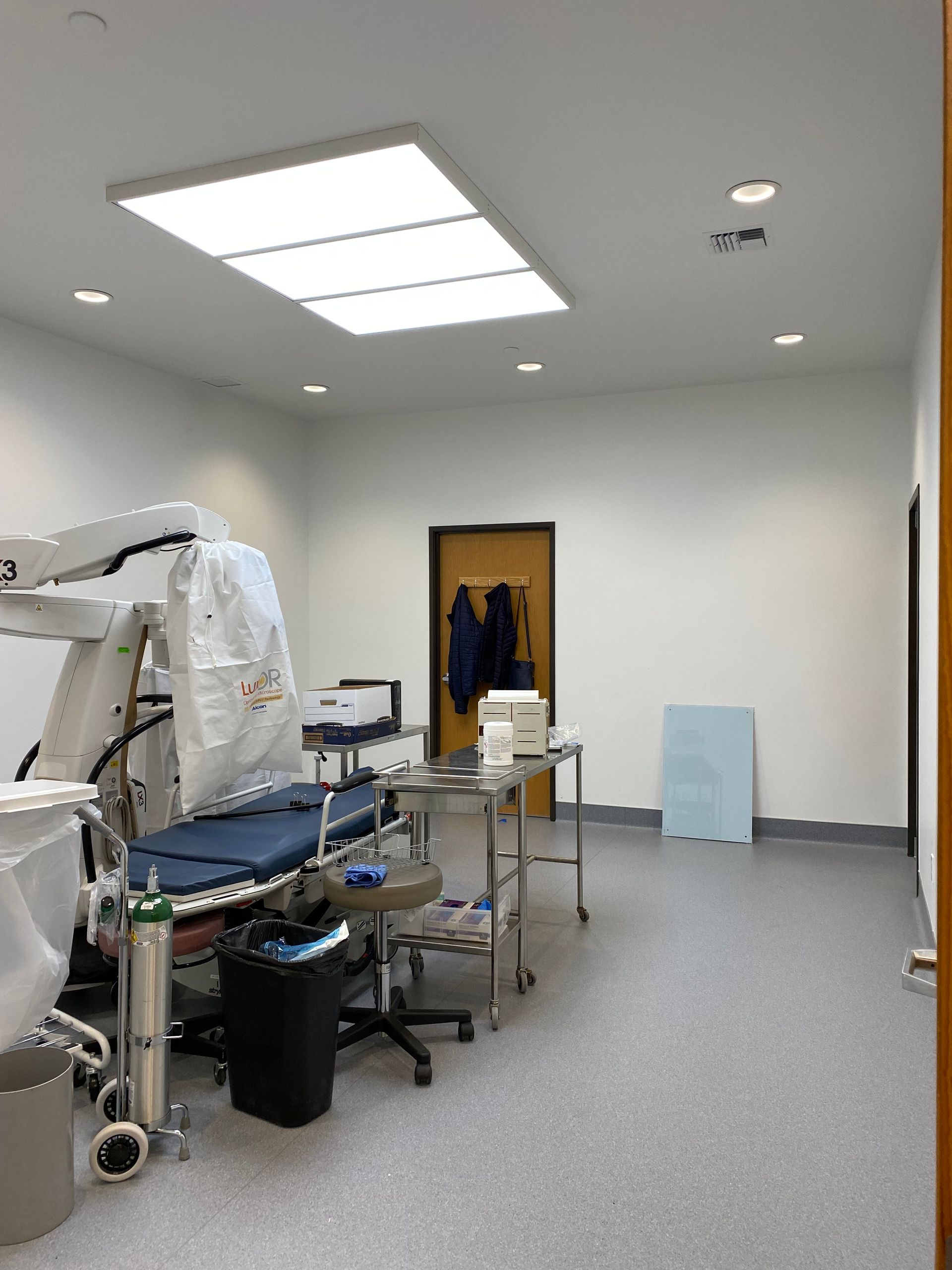 Medical examination room with equipment, medical bed, and door. White walls, gray floor, bright ceiling lights.