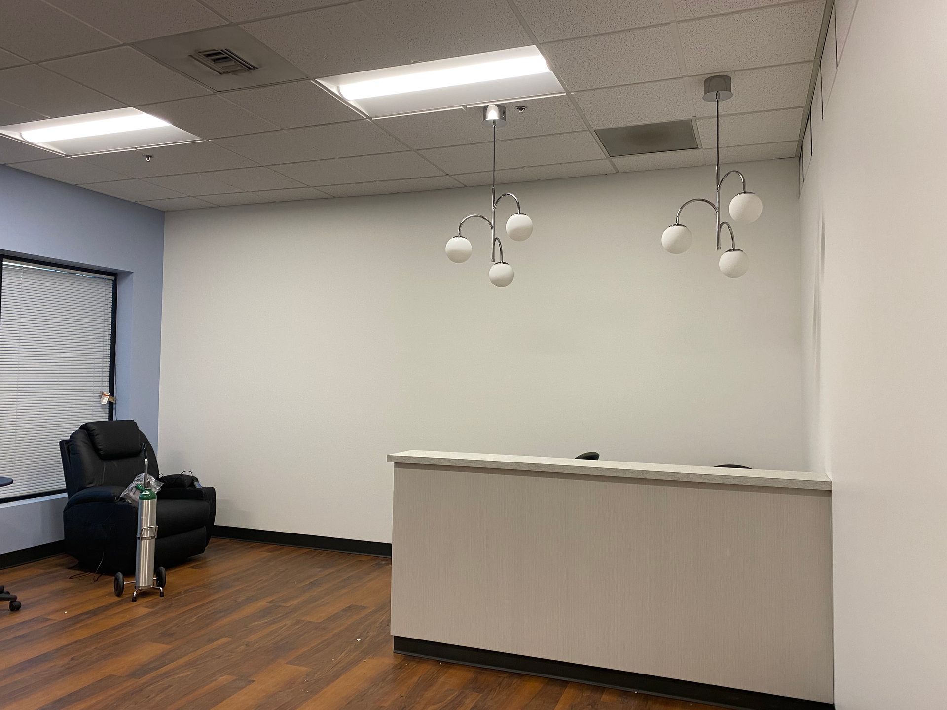 Reception area: desk, chandelier lights, chair, window with blinds, wood floor, white walls.
