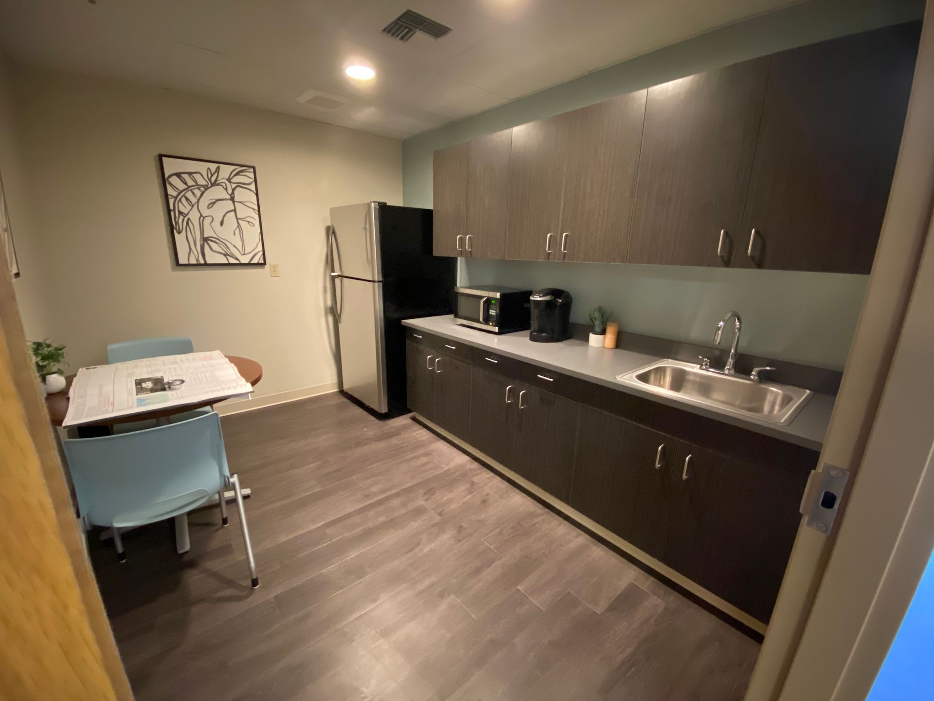 Small office kitchen with dark cabinetry, stainless steel fridge, table, chair, artwork.