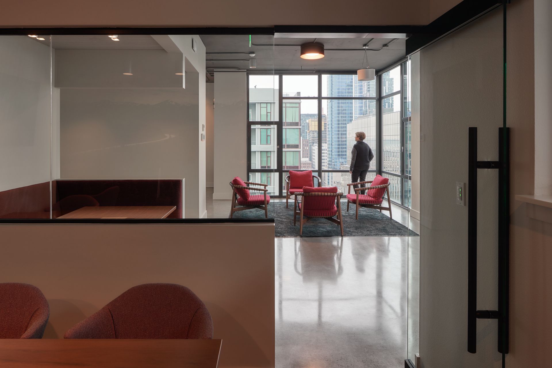 A person looking out a large window in a modern room with red chairs and a gray rug.