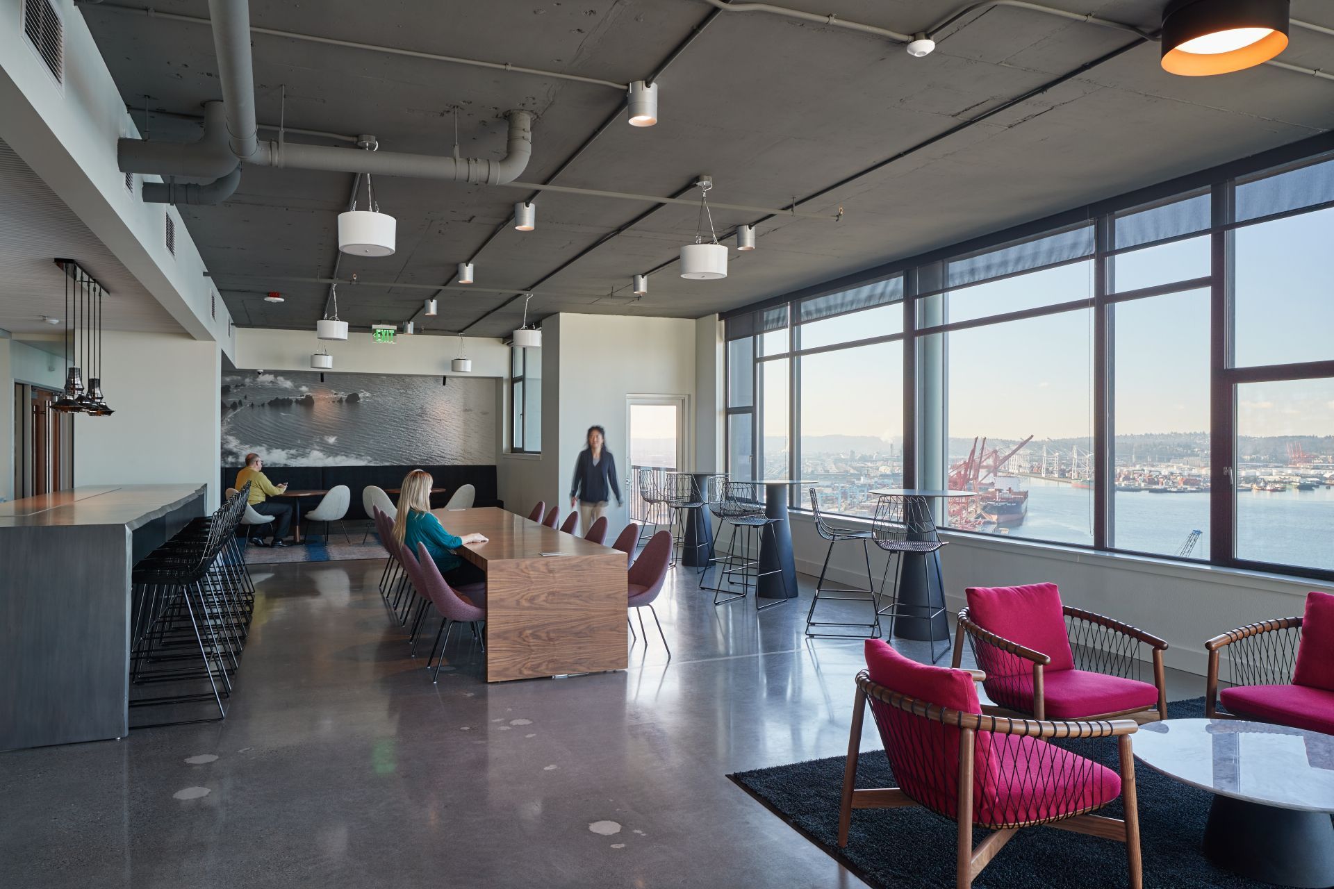 Spacious modern lounge with cityscape view. People seated at tables, red chairs, polished concrete floor, and large windows.