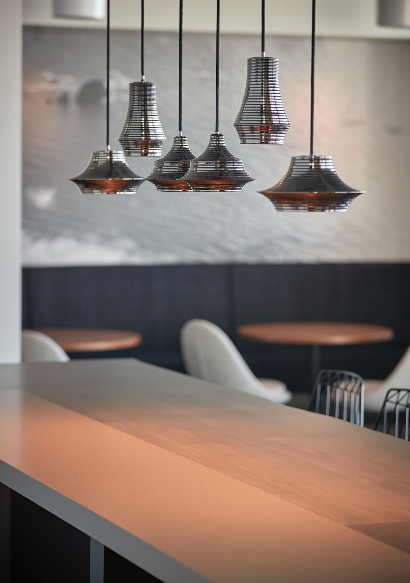 A row of hanging, silver pendant lights illuminates a long table in a modern cafe.