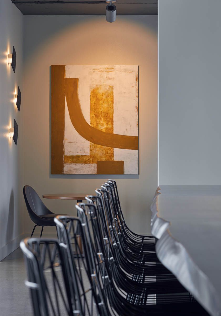 A room with an abstract painting in gold and white, black chairs, and sconce lighting.