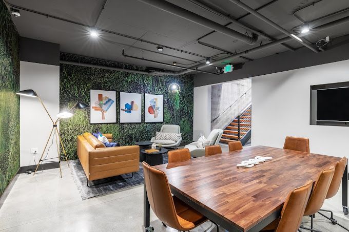 Modern lounge with a large wooden table, chairs, a couch, art, and a faux-grass wall.