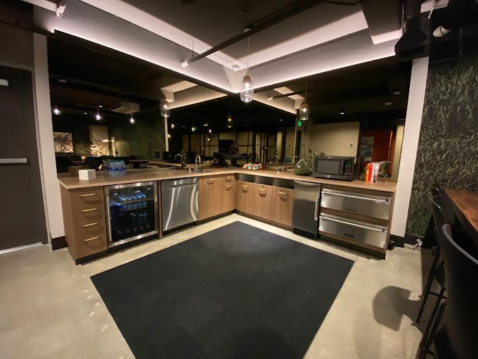 L-shaped office kitchen with wooden cabinets, stainless steel appliances, and a black rug.