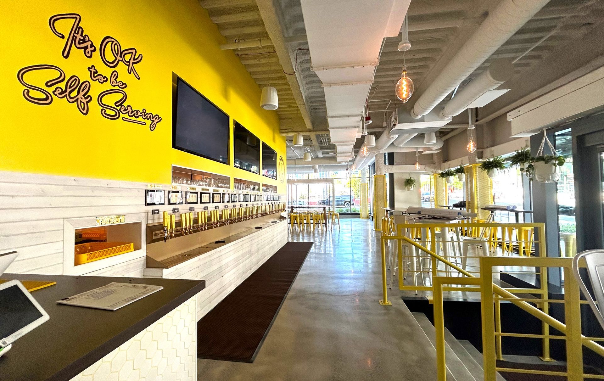 Bright yellow-walled bar with beer taps and a seating area; concrete floors, large windows, and overhead lighting.