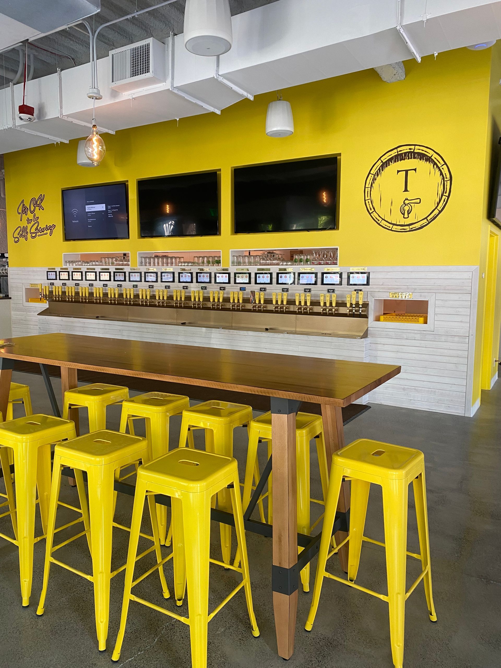 Yellow bar stools and wall with beer taps. Three TVs above the taps.