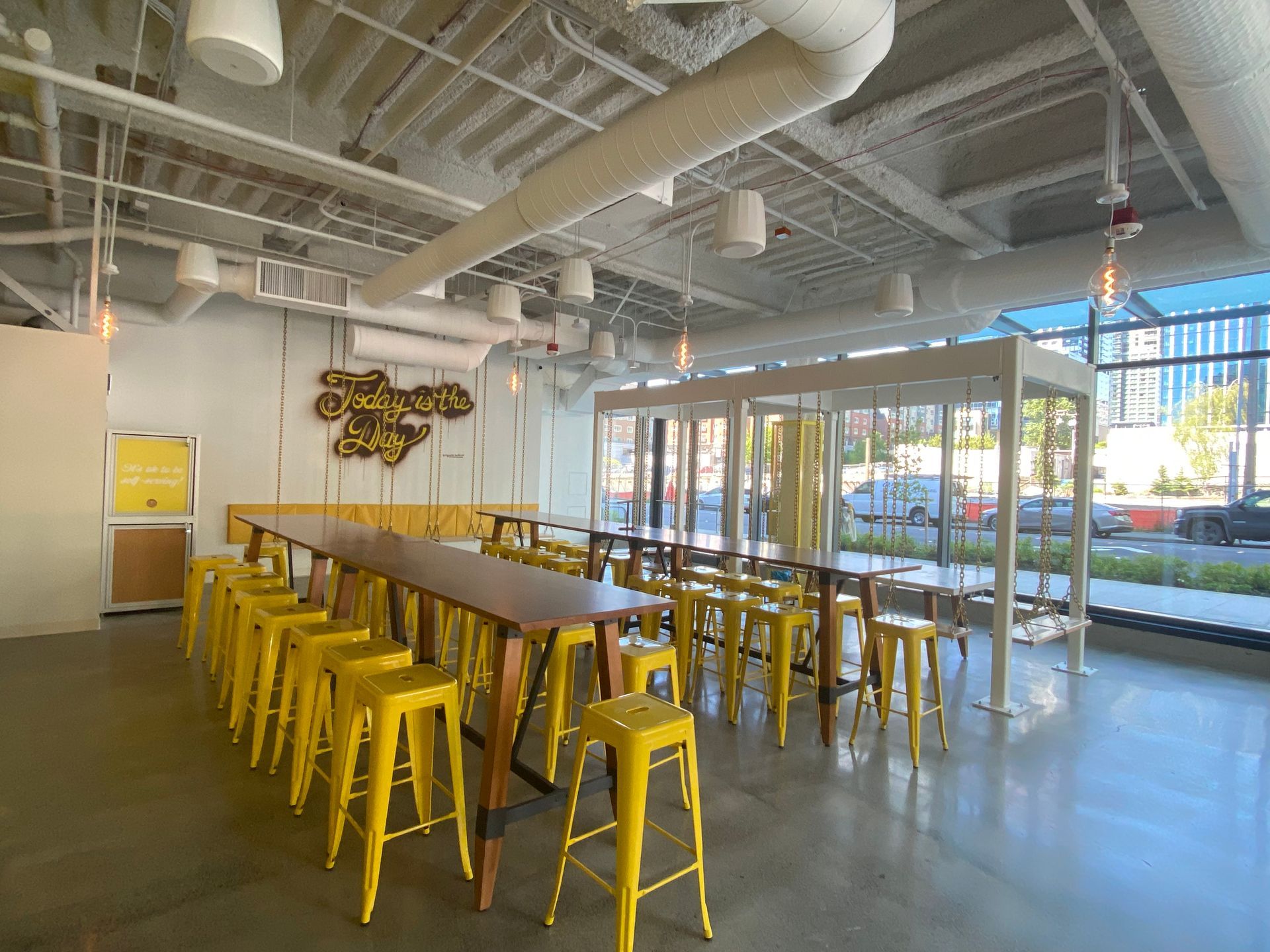 Interior of a brightly lit cafe with yellow stools and a long wooden table. Glass enclosed seating area visible.