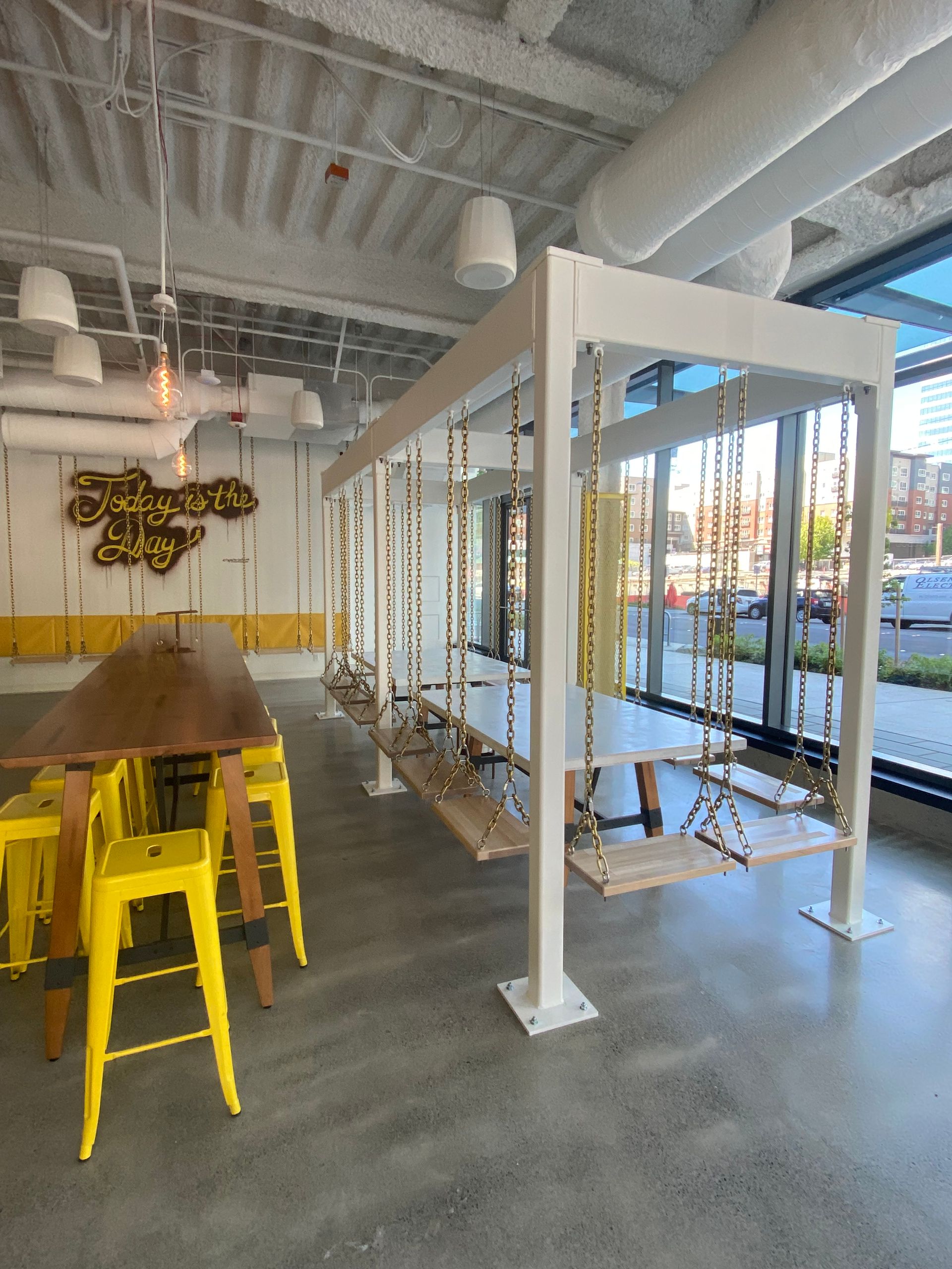 Interior with swings, a long table, and yellow stools. A sign hangs on the wall.