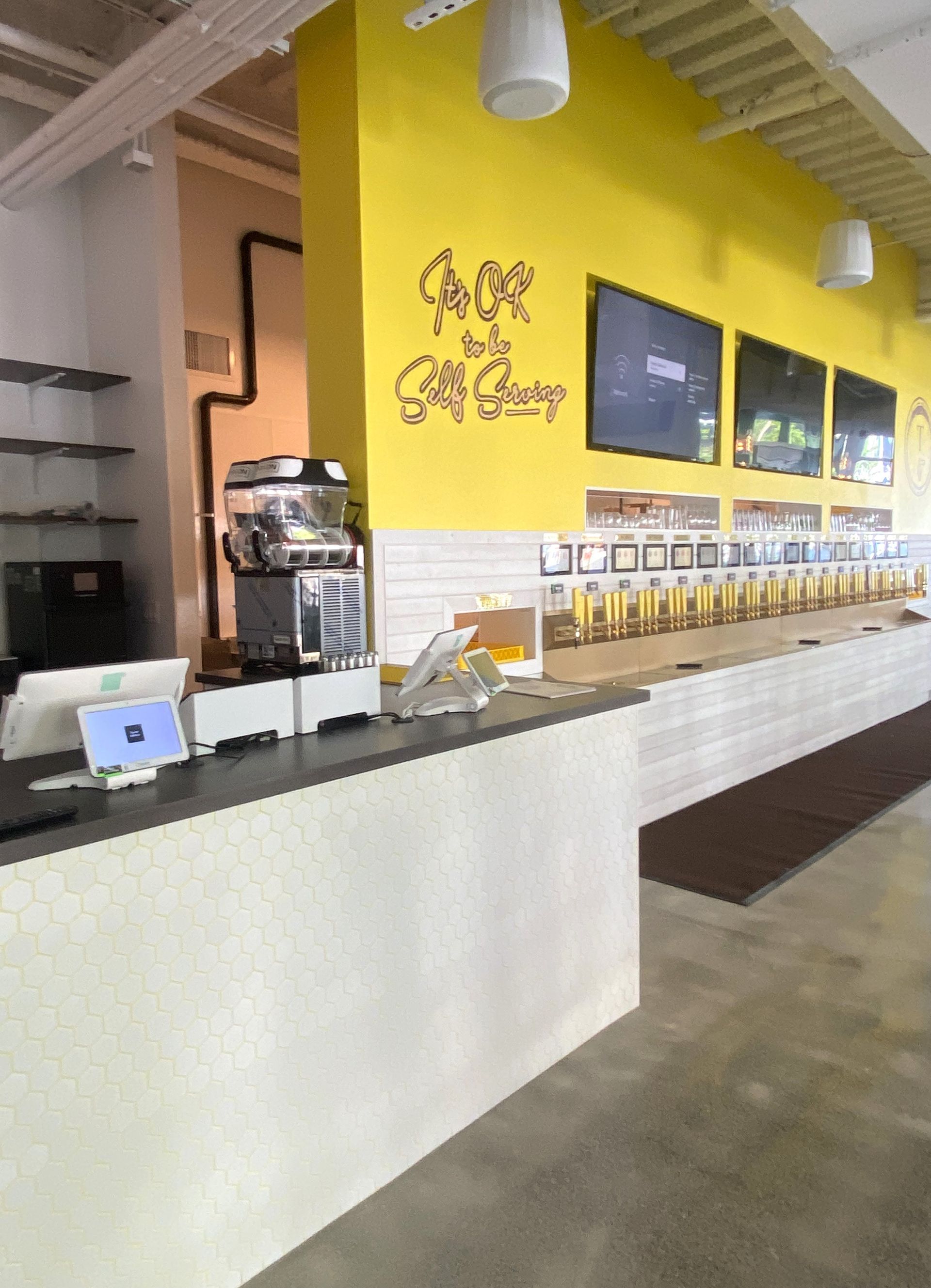 Bar with yellow wall, tap handles, and multiple screens. White and gray counters.