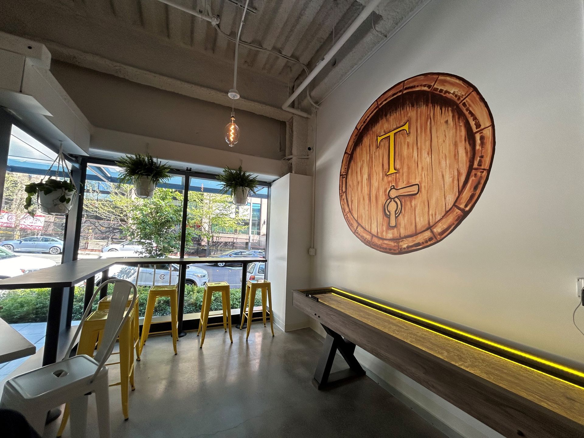 Interior of a bar with a shuffleboard table, a barrel logo, and seating near a window overlooking a street.