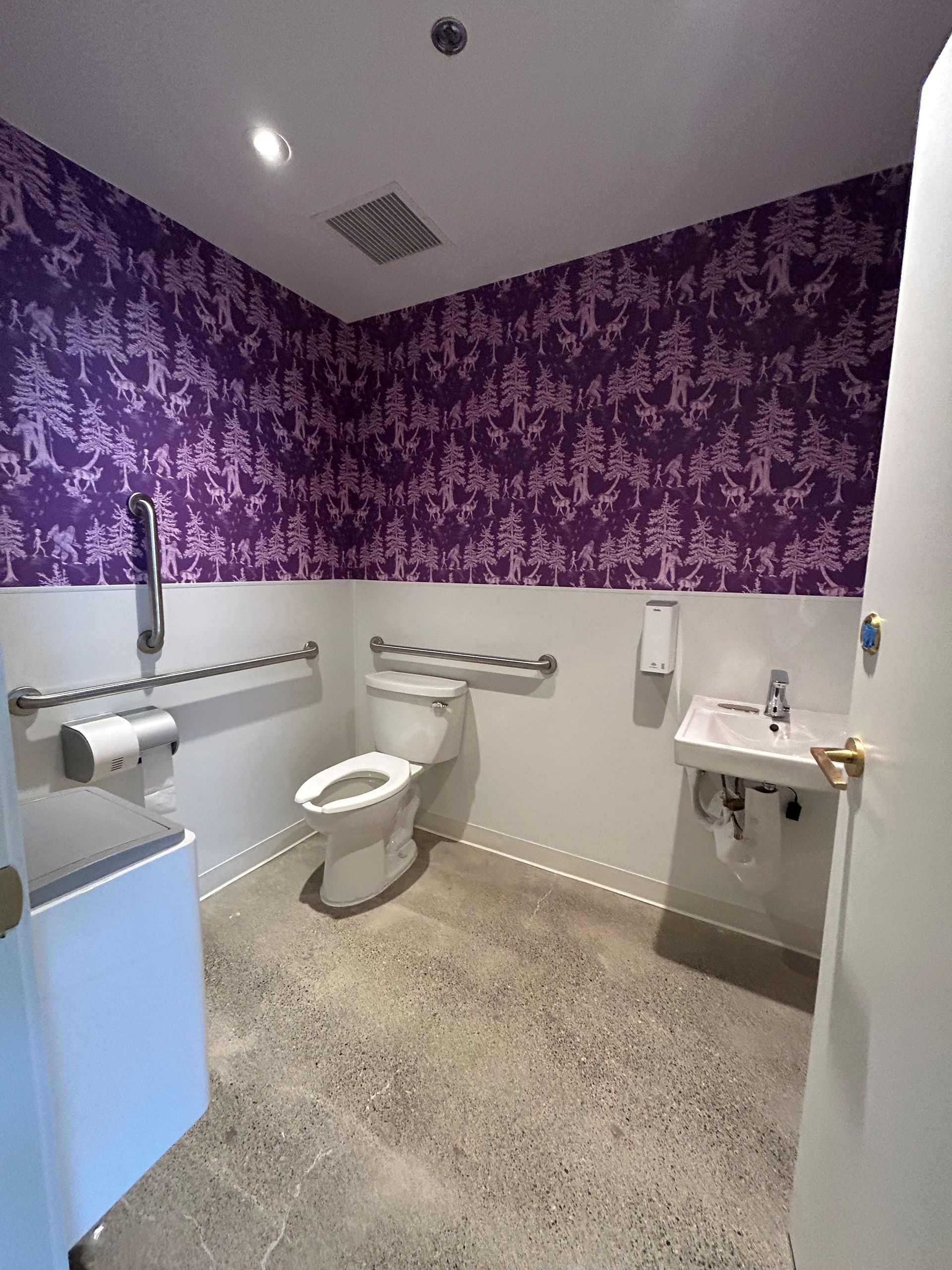Accessible restroom with purple forest wallpaper, toilet, sink, grab bars, and light gray floor.