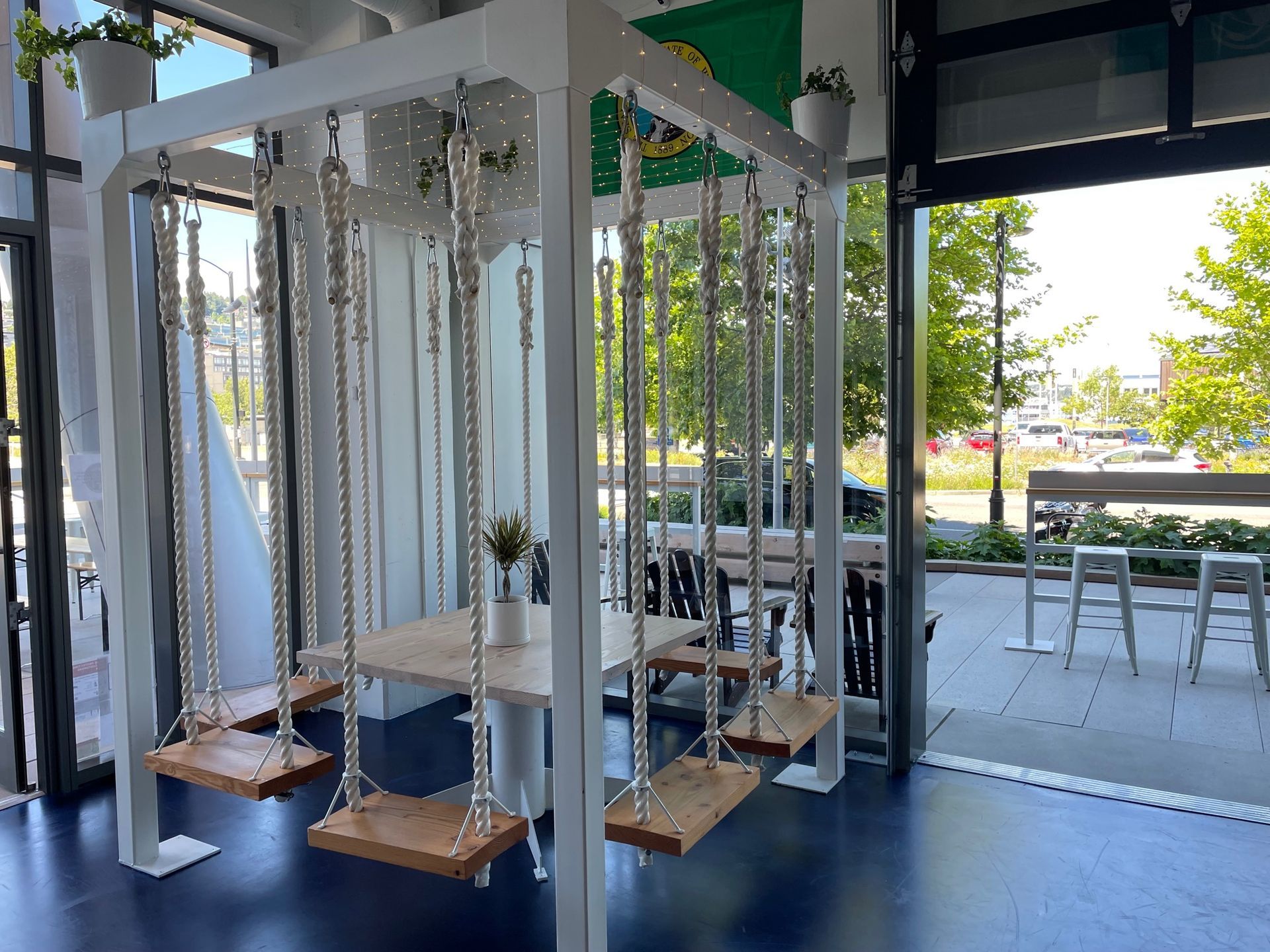A whimsical indoor dining area with a swing table. White frame holds swings around a light wood table, open to outdoor seating.