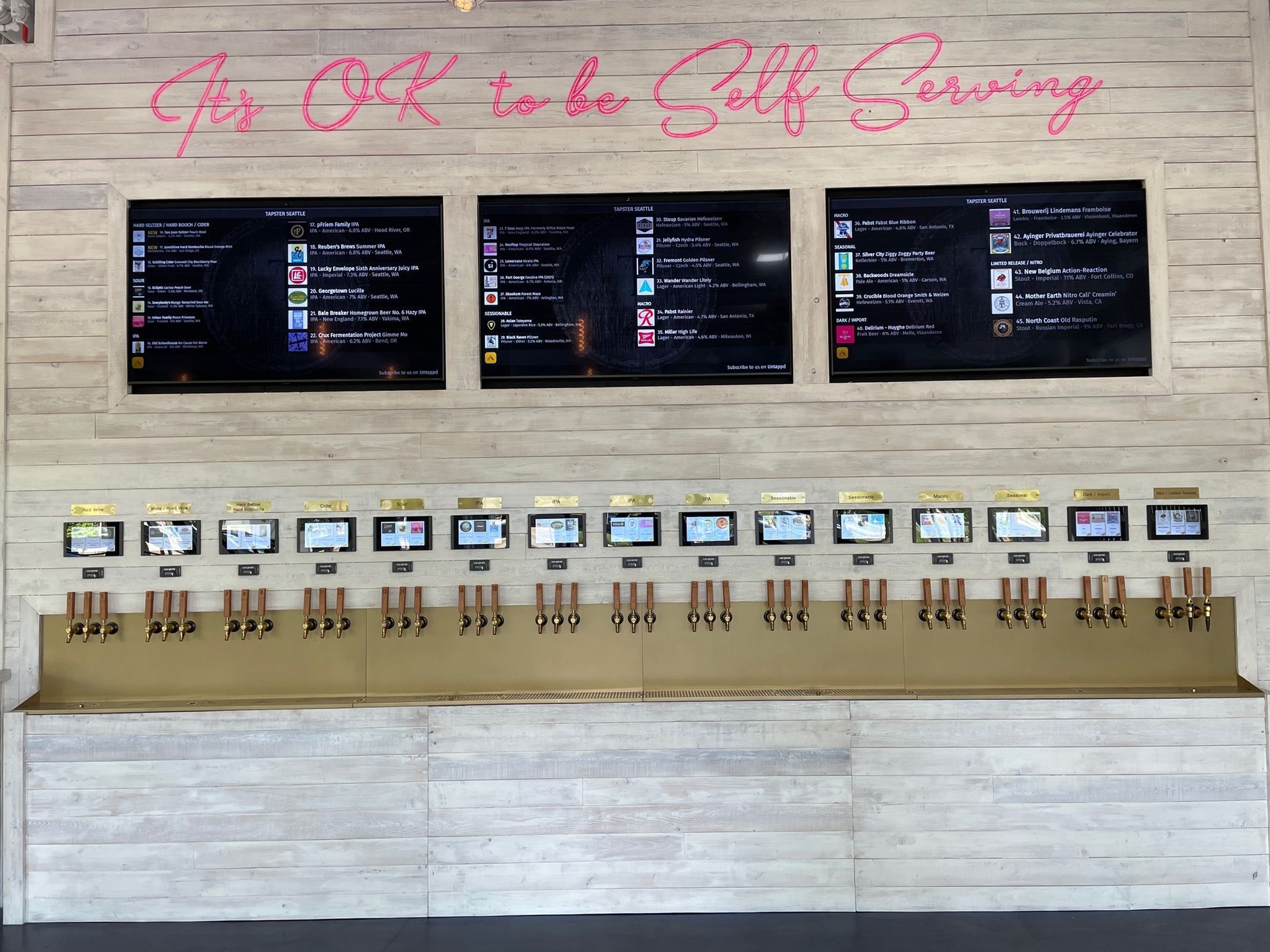 A self-serve taproom. Taps line a wall under three screens displaying drink options. Neon sign: 