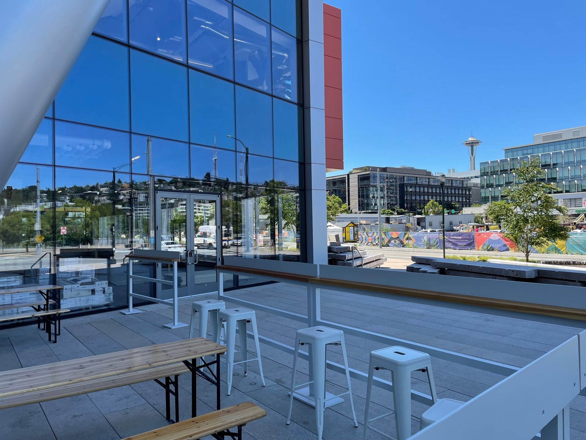 Outdoor patio with tables and stools, overlooking city buildings, including the Space Needle, on a sunny day.