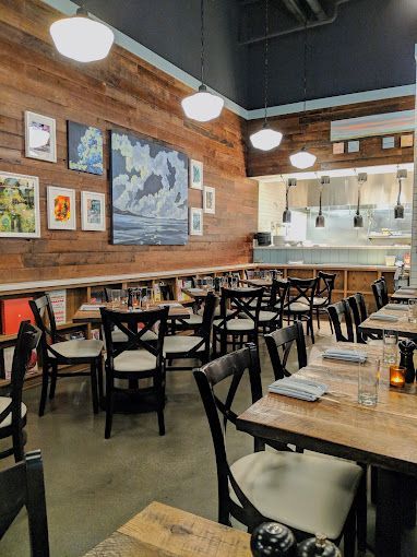 Restaurant interior with wood paneling, tables, chairs, and artwork; a kitchen is visible in the background.