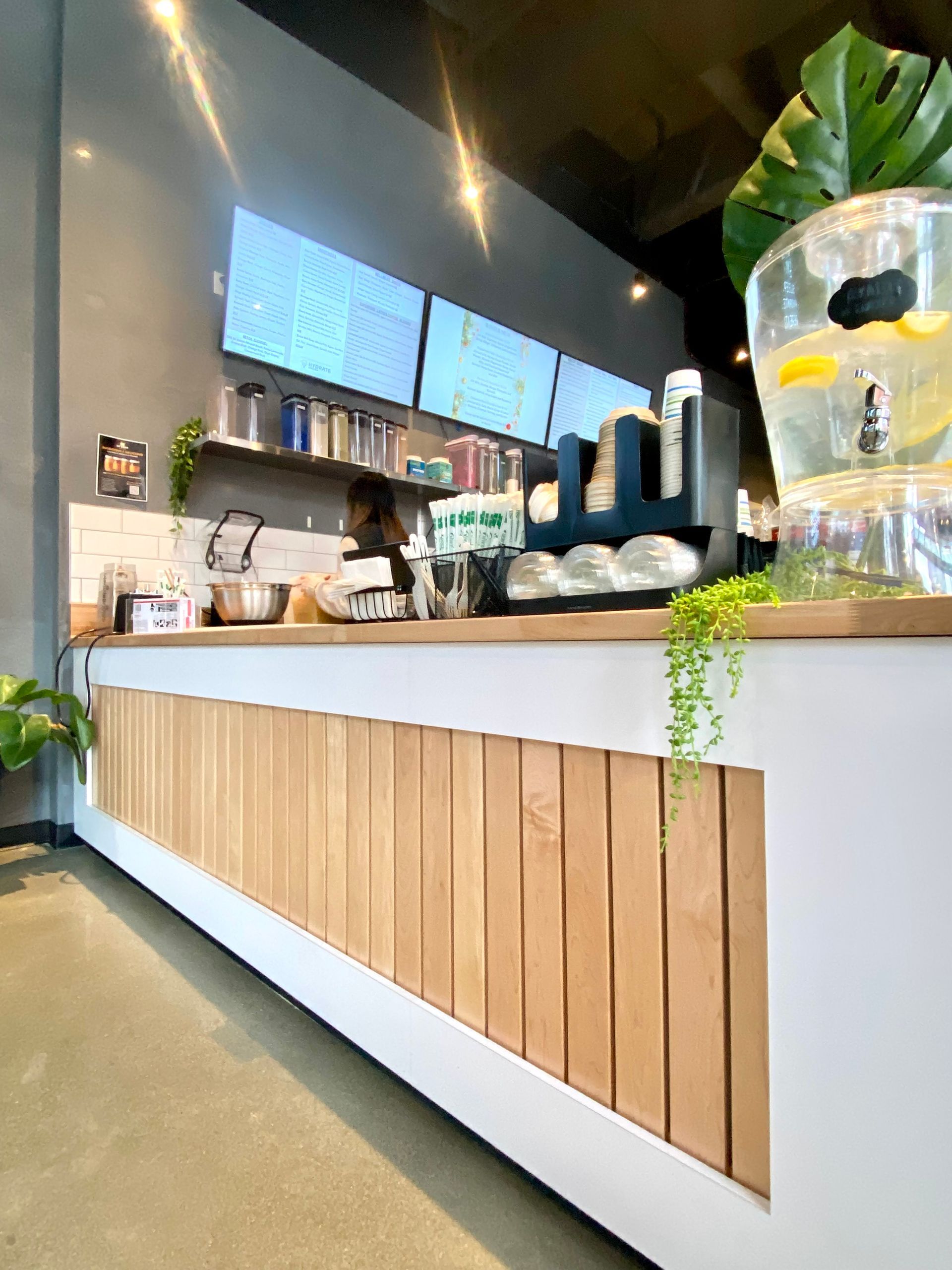 Counter in a cafe with wood paneling. Menu boards, drink dispensers, and a person are behind the counter.