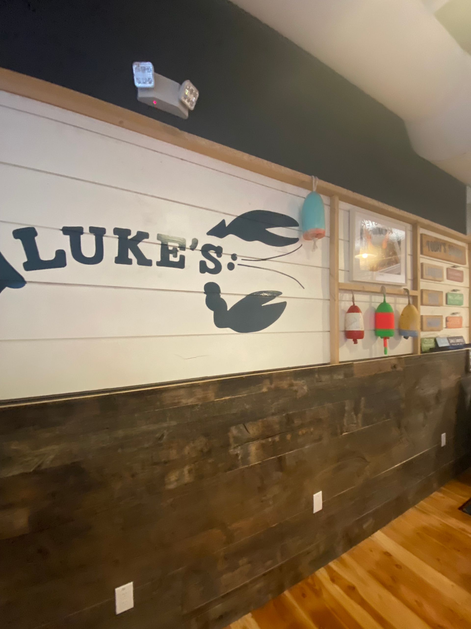 Wall at Luke's Lobster restaurant with logo, lobster claws, and decorative buoys. Dark wood paneling below.