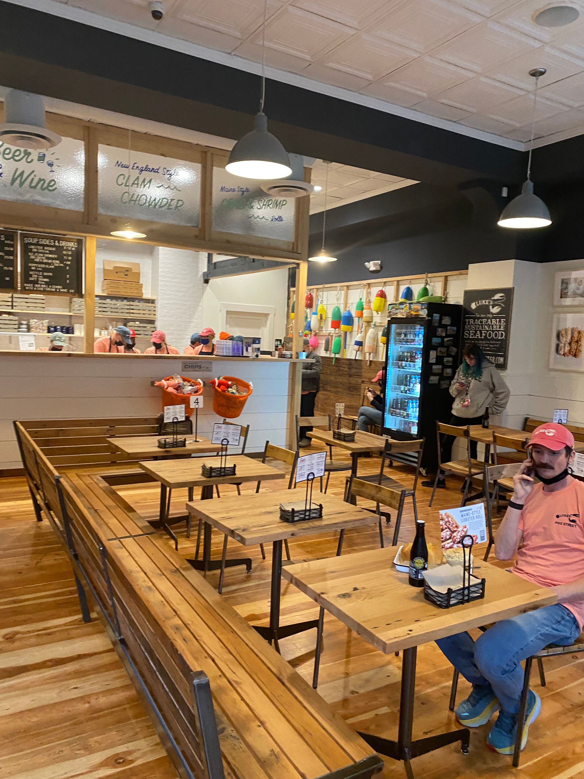 Interior of a restaurant: wooden tables and benches, overhead lights, a person on the phone, and a counter with menu boards.