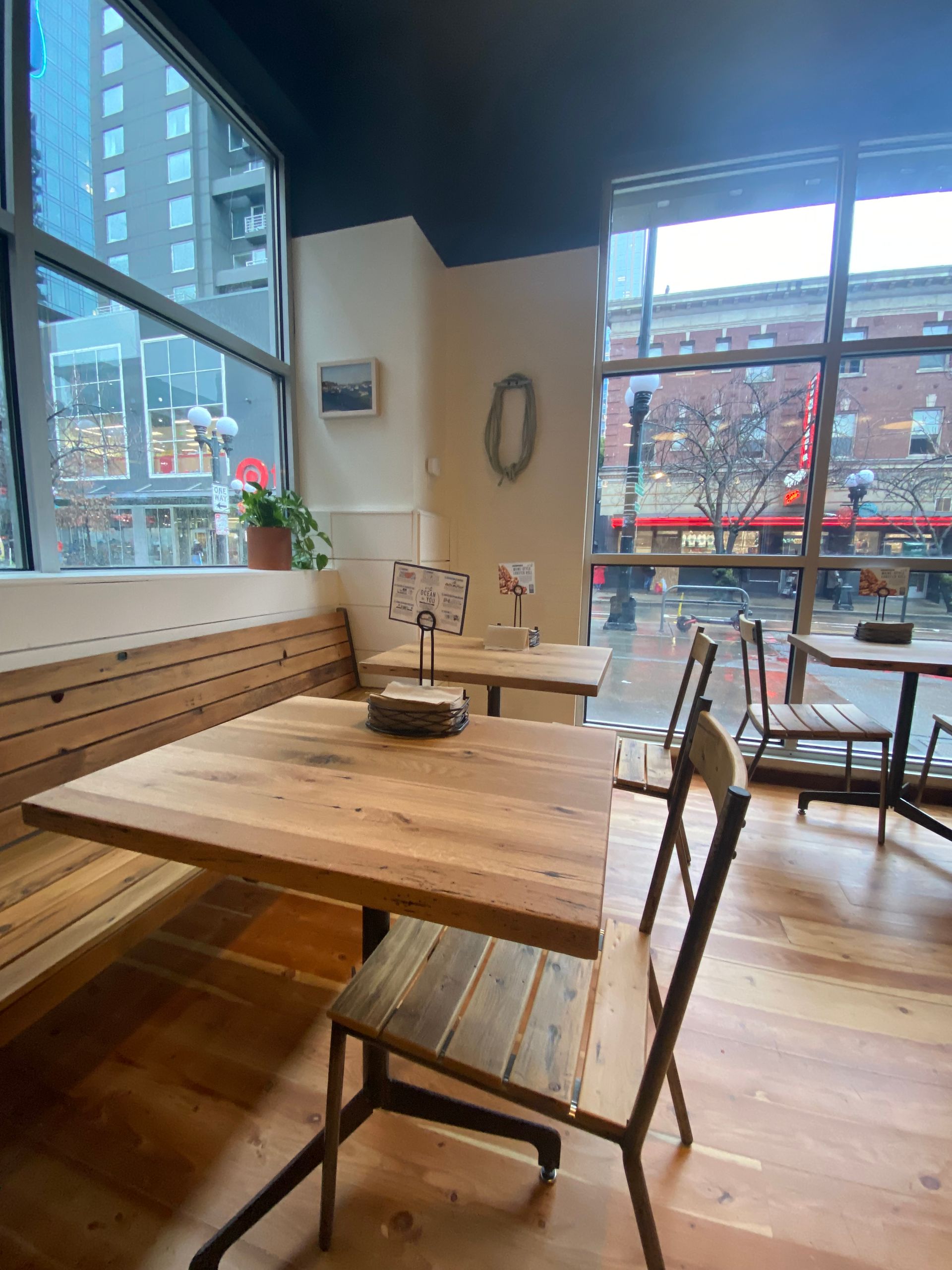 Cafe interior: wood tables and chairs, large windows with city view, light wood floors, and neutral walls.