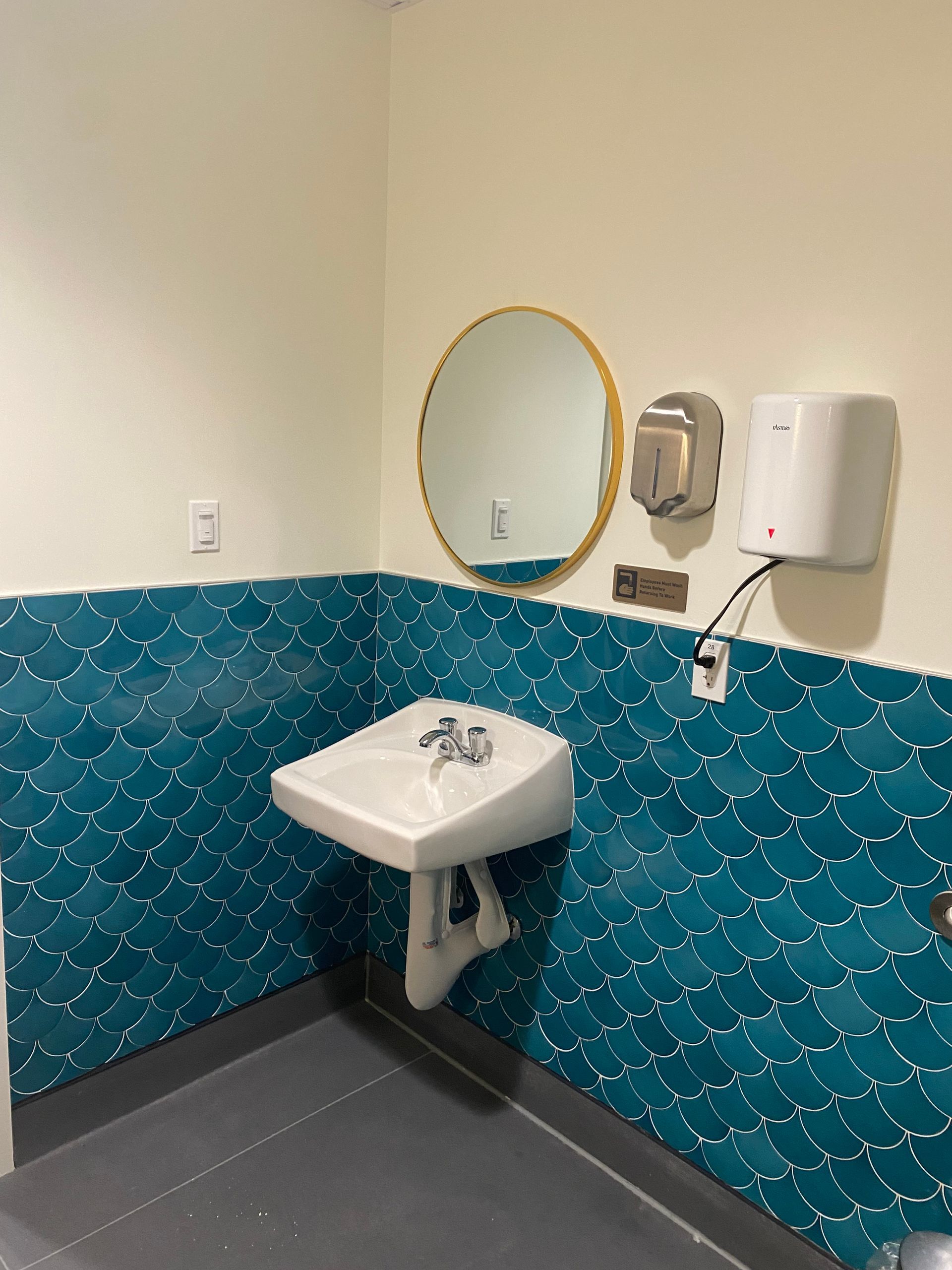 Bathroom with a white sink, teal tile backsplash, round mirror, and hand dryer.