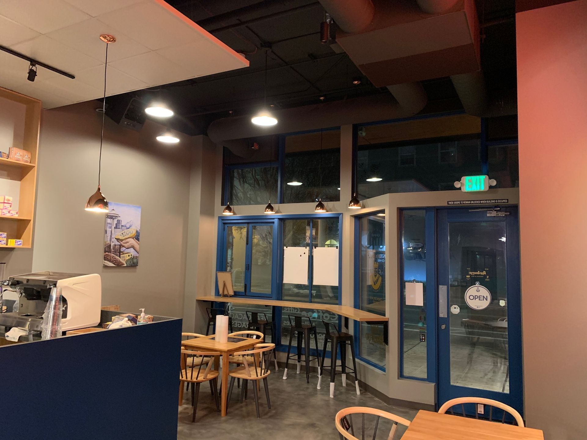 Interior of a cafe with tables, chairs, bar seating, windows, and hanging lights.