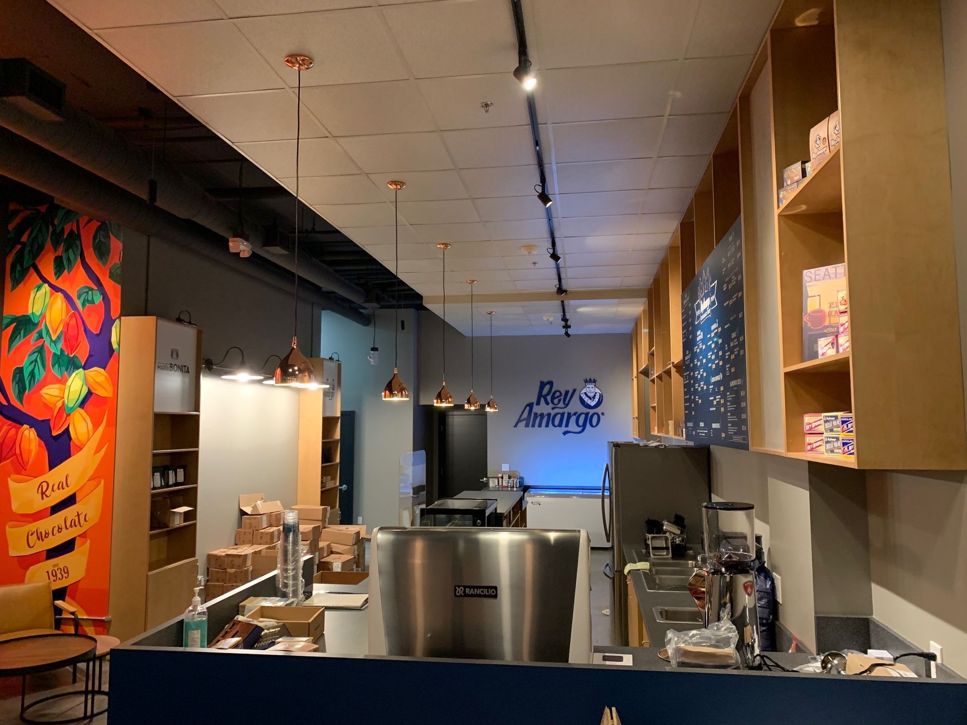Interior of a cafe with a counter, shelves, and a colorful mural. Copper-colored lights hang from the ceiling.