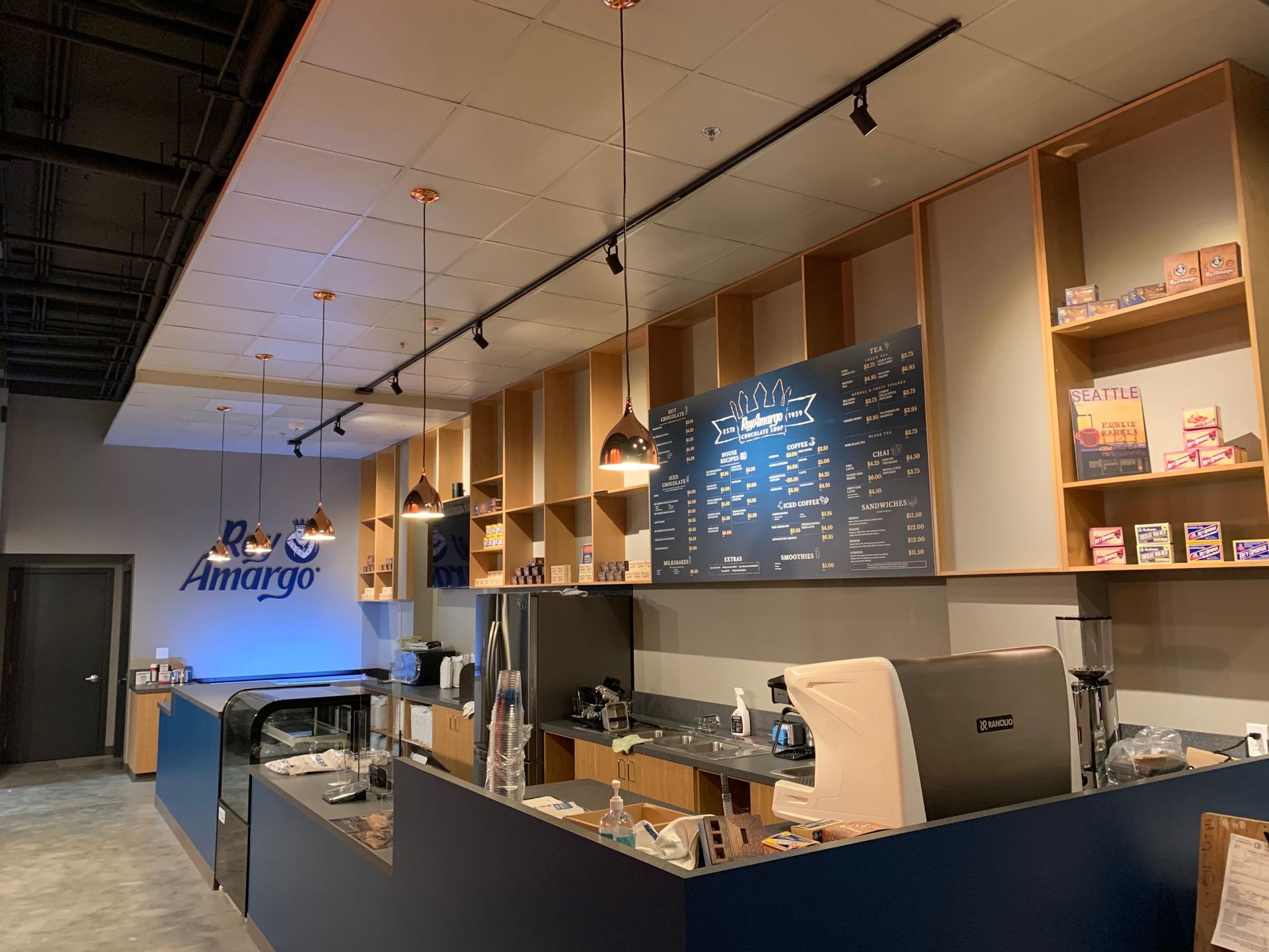 Coffee shop interior with bar, menu board, and shelving filled with products. Dark blue and wood tones.