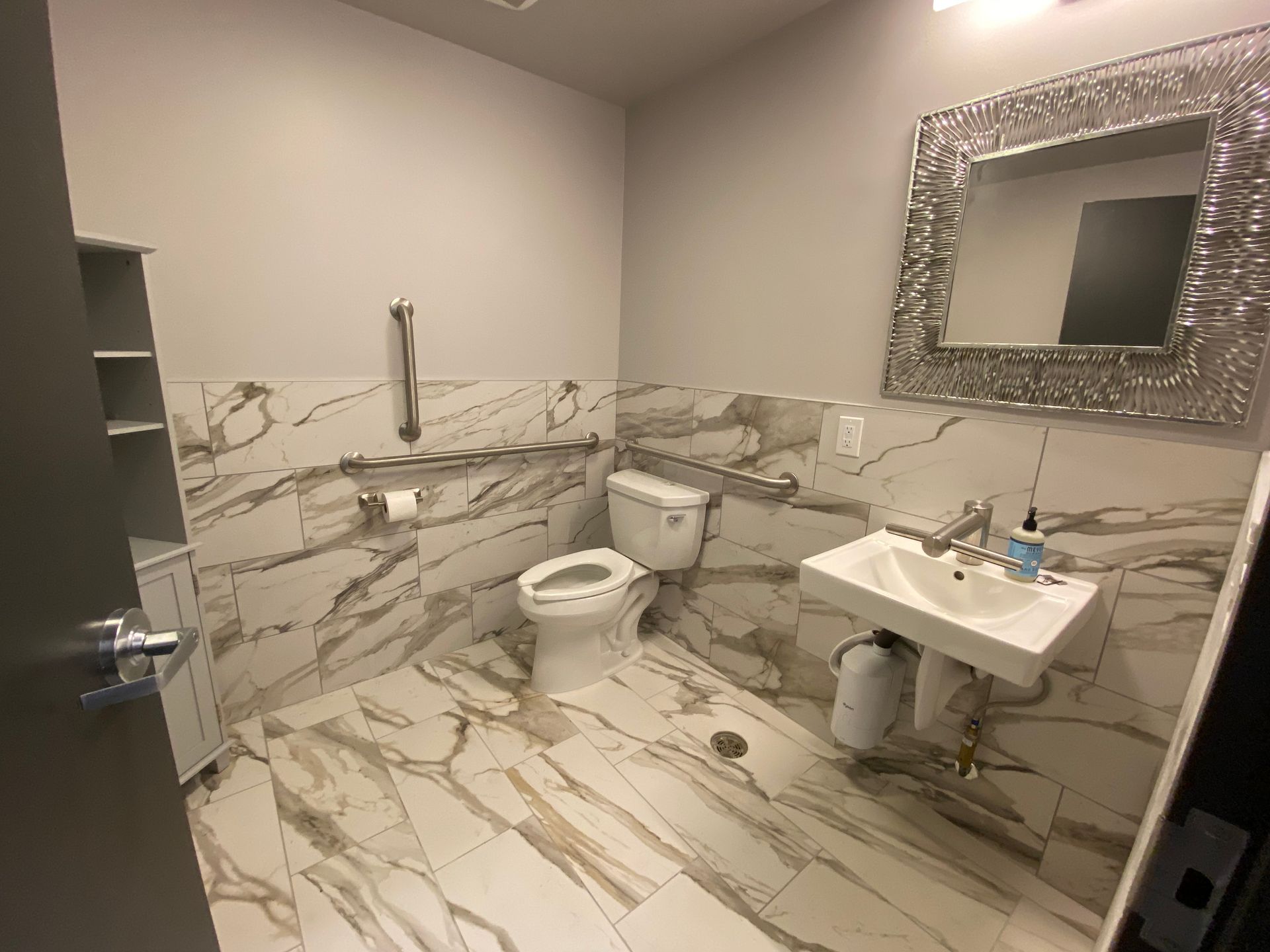 Bathroom with marble-look tile, white sink and toilet, silver framed mirror, grab bar, and small cabinet.