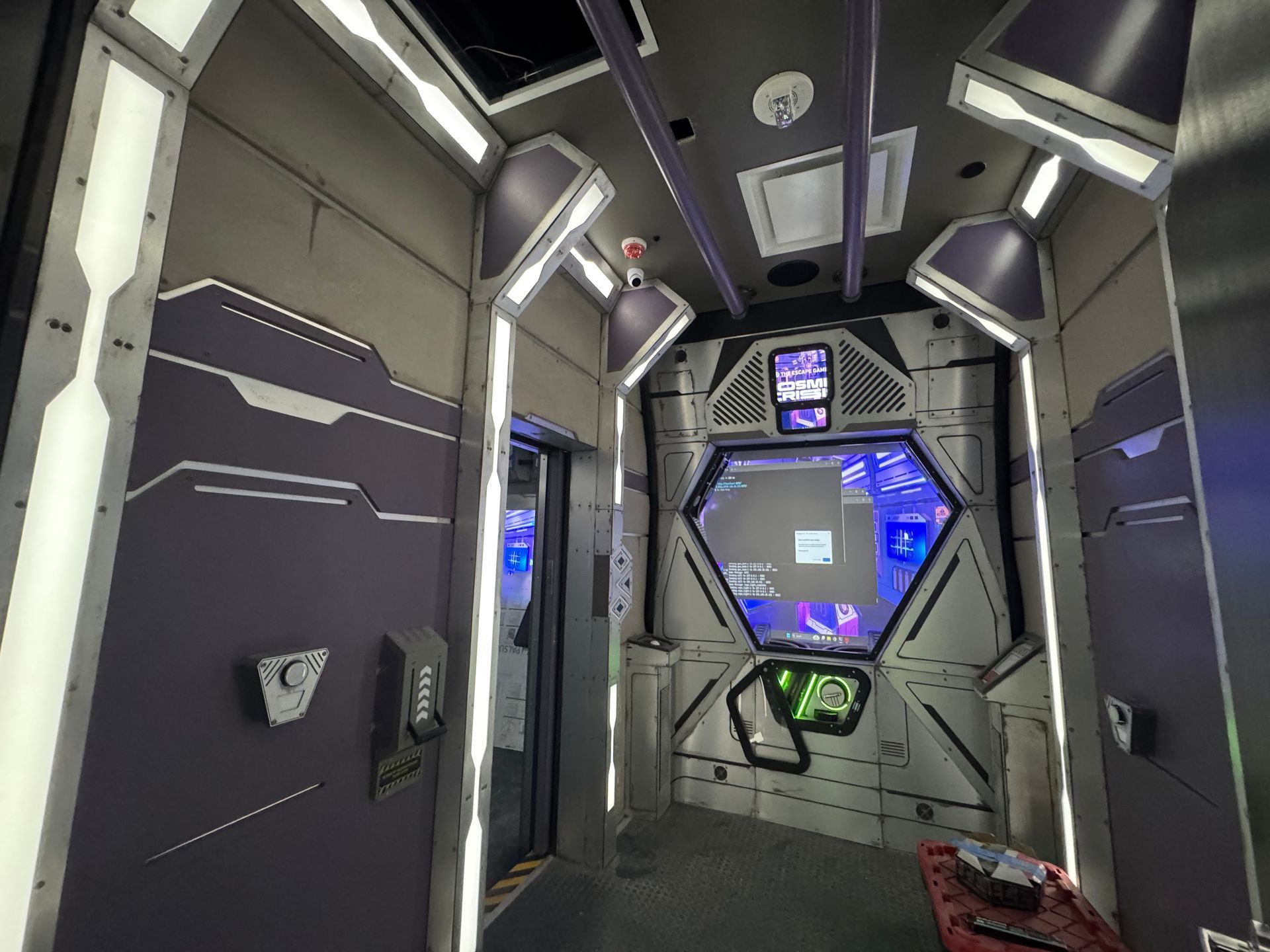 Interior of a futuristic-looking elevator with metallic gray walls, a control panel, and hexagon window with blue lighting.