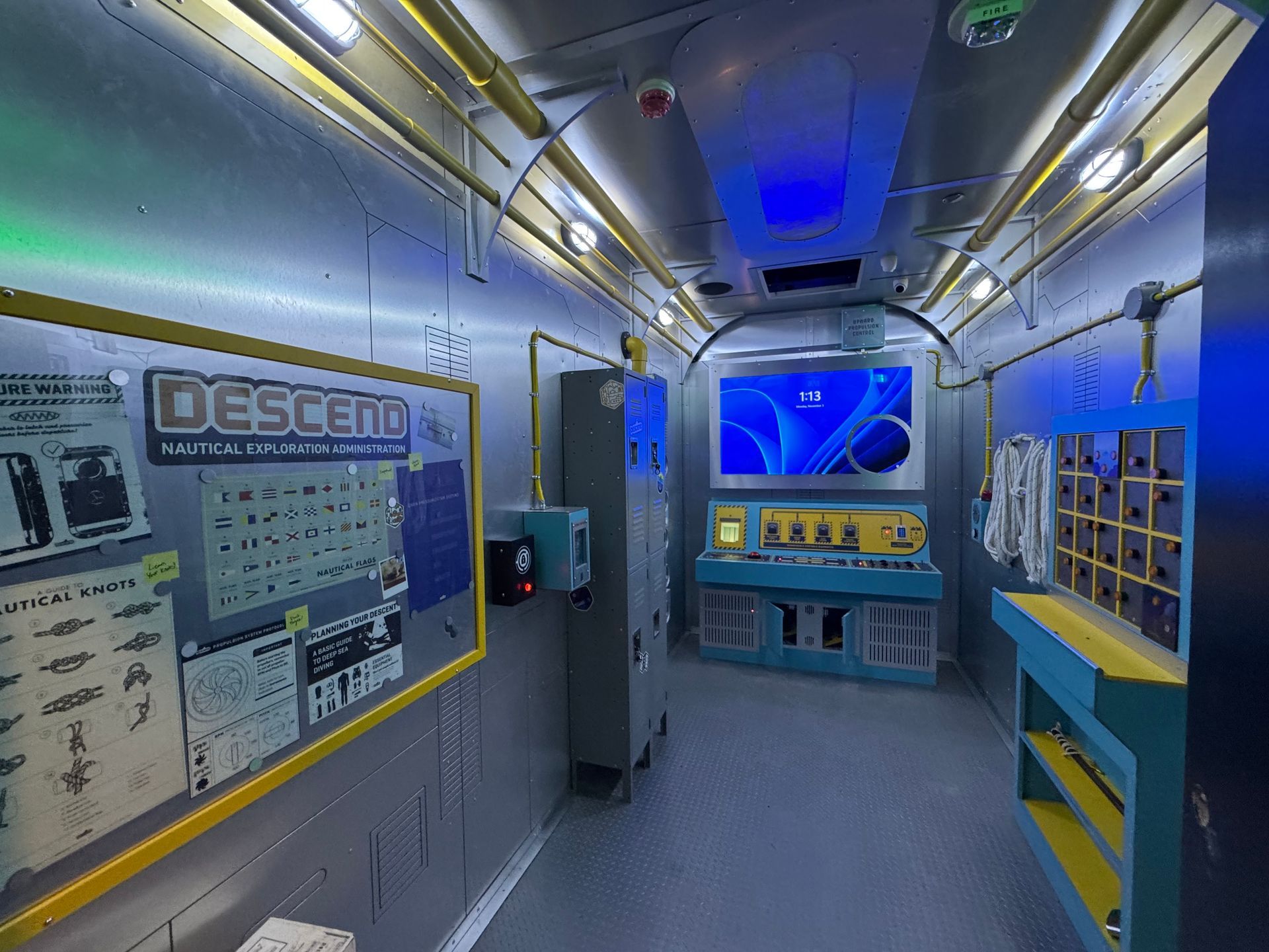 Interior of a submarine-like room with control panels, a screen showing a blue image, and various tools.