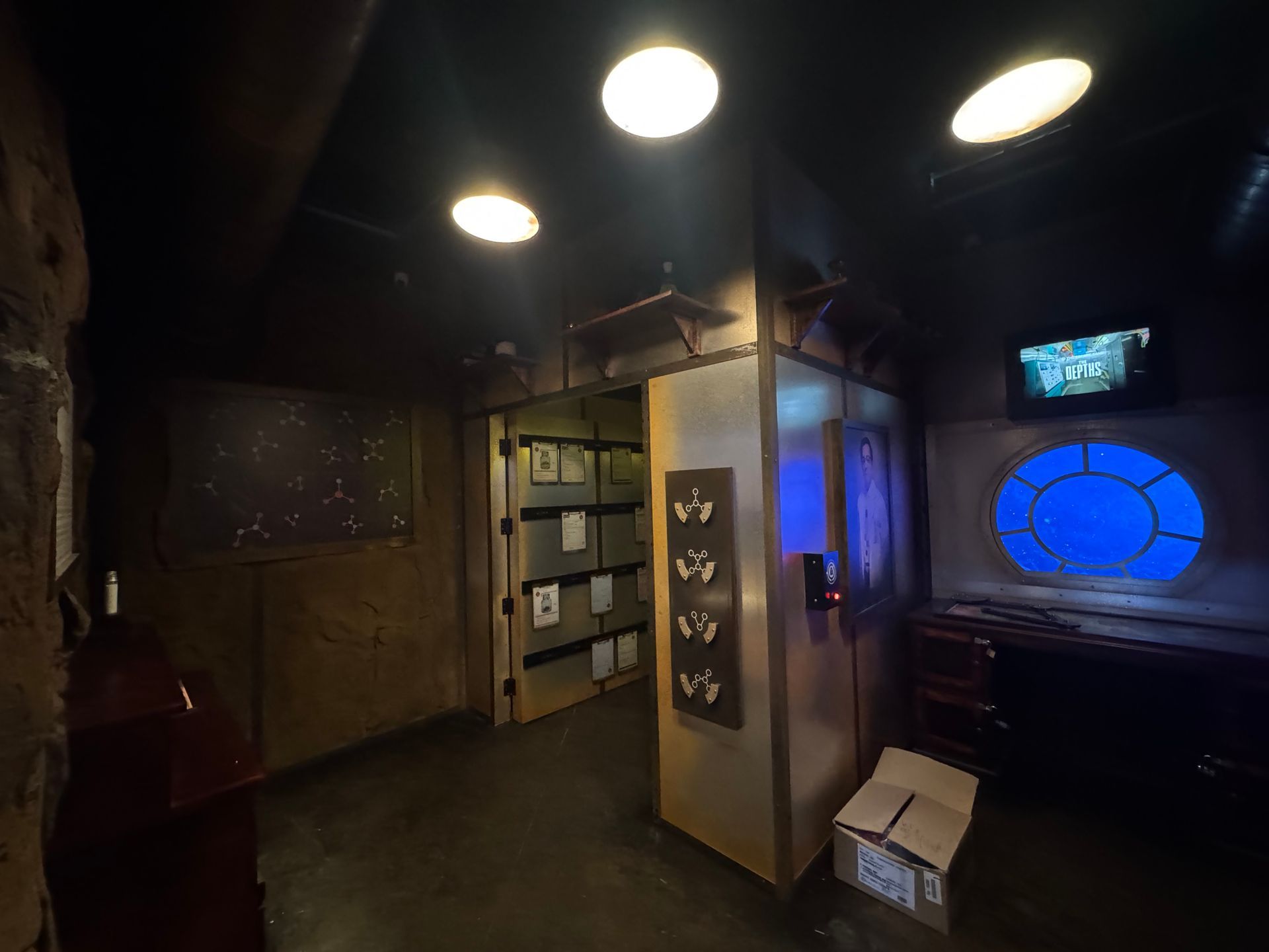 Interior of a dimly lit submarine control room. Features a control panel, desk, porthole, and shelving.