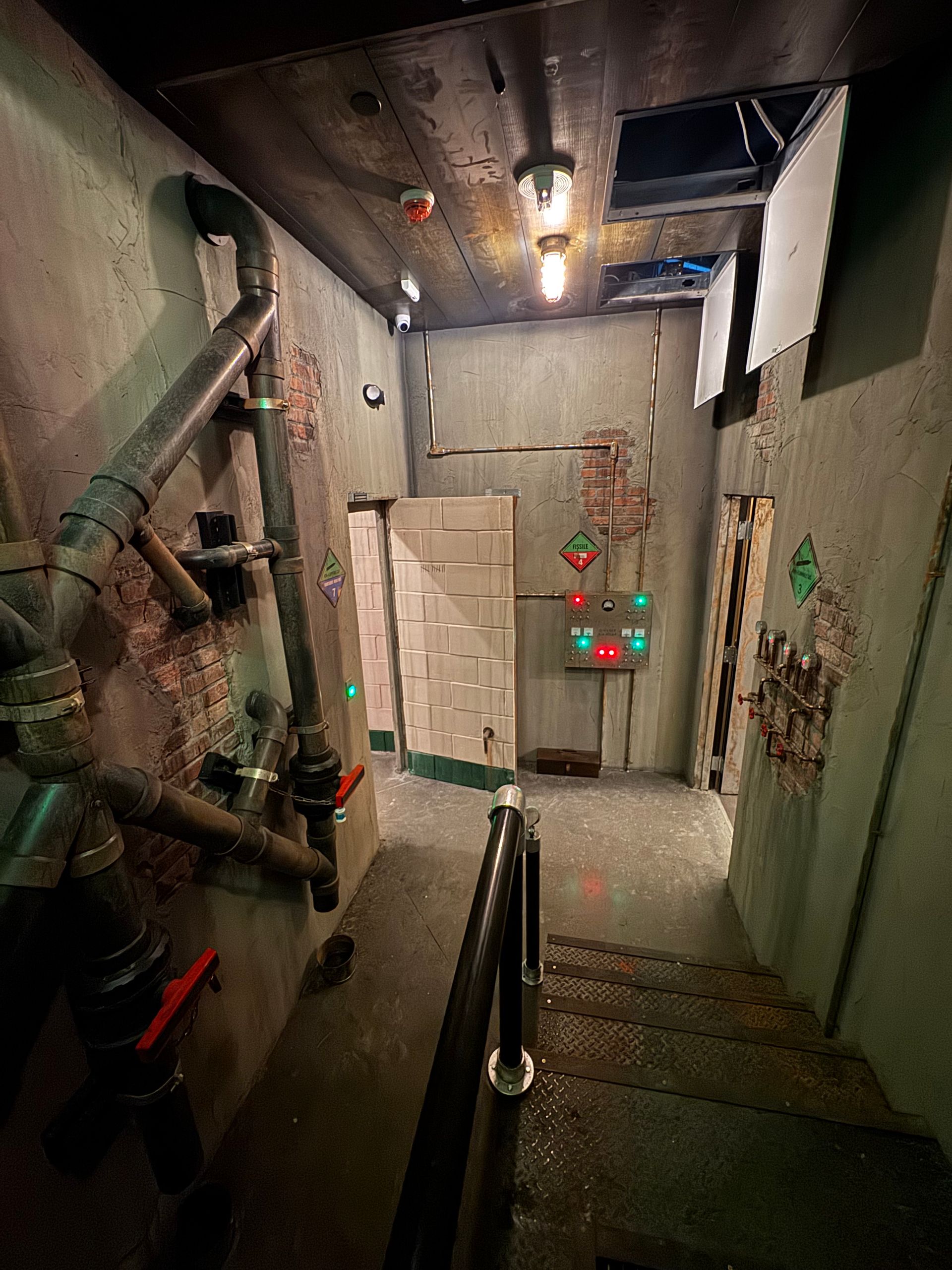 A dimly lit, industrial-style hallway with stairs, pipes, and control panel.