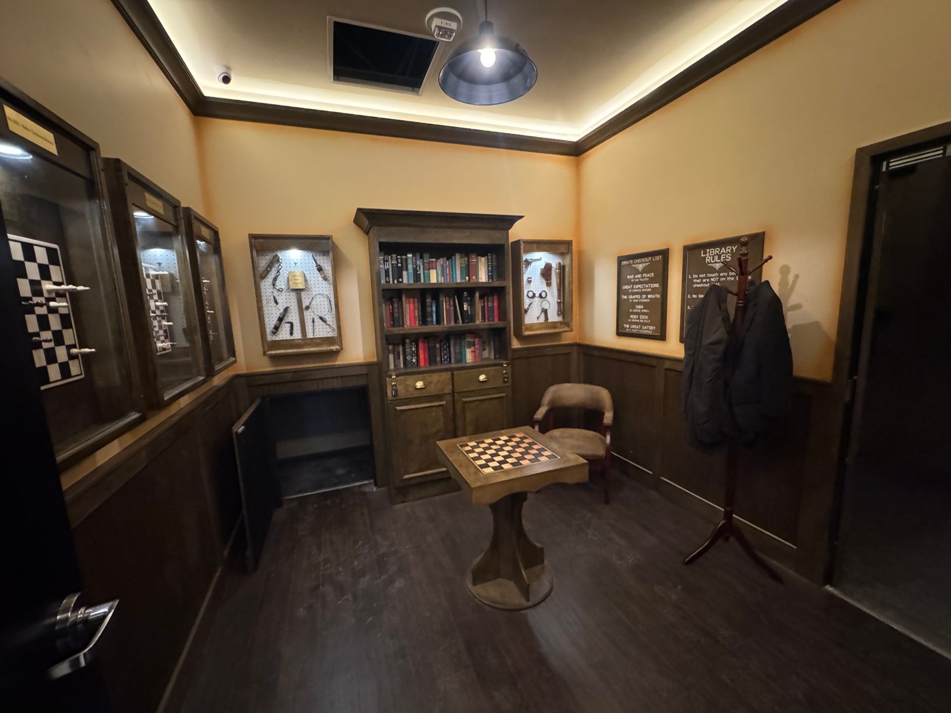 A dimly lit room with a bookcase, chessboard, display cases, and a coat rack; likely an escape room.