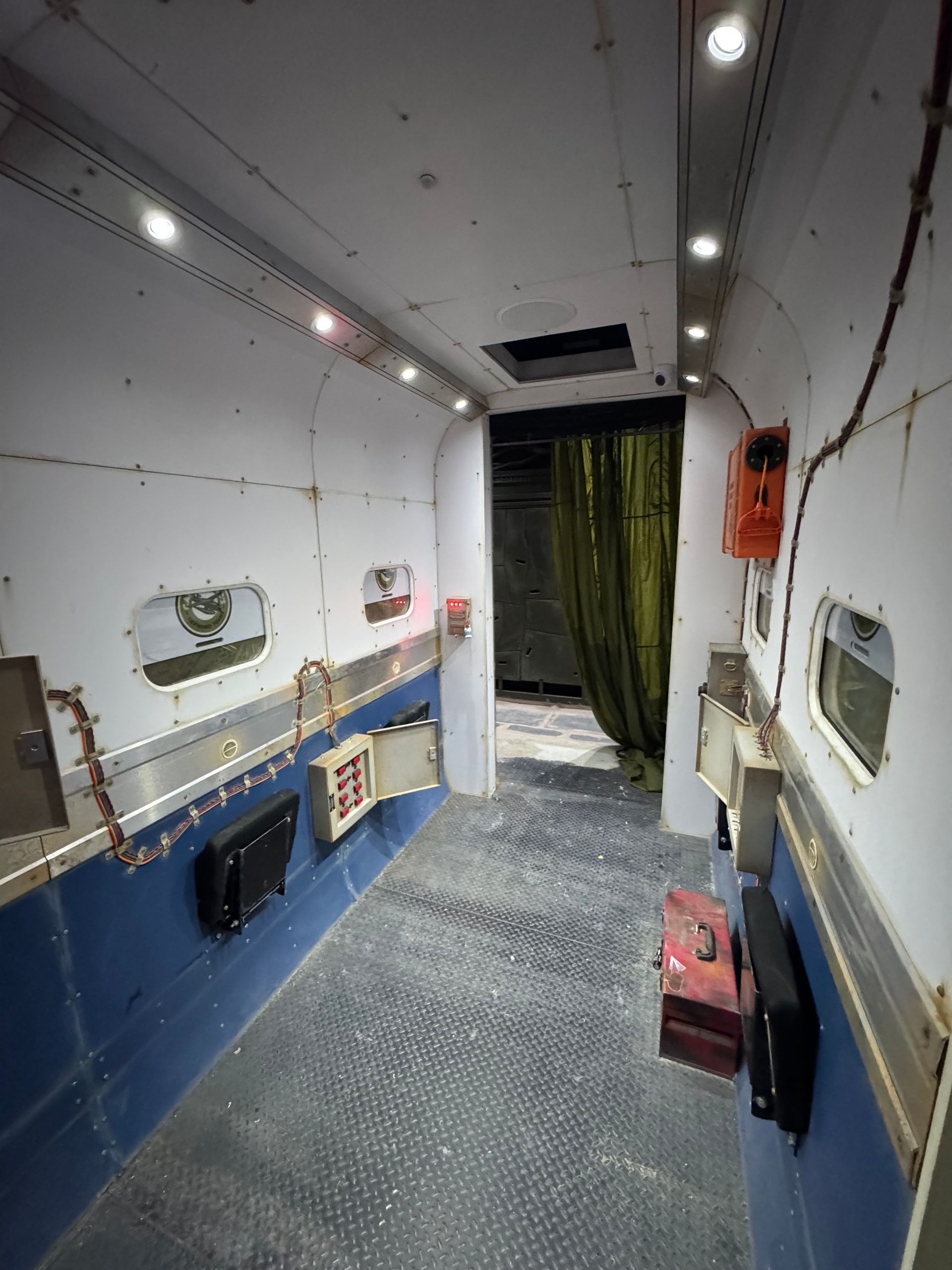 Narrow interior hallway, white walls with rectangular windows, leading to a dark opening with green curtain.