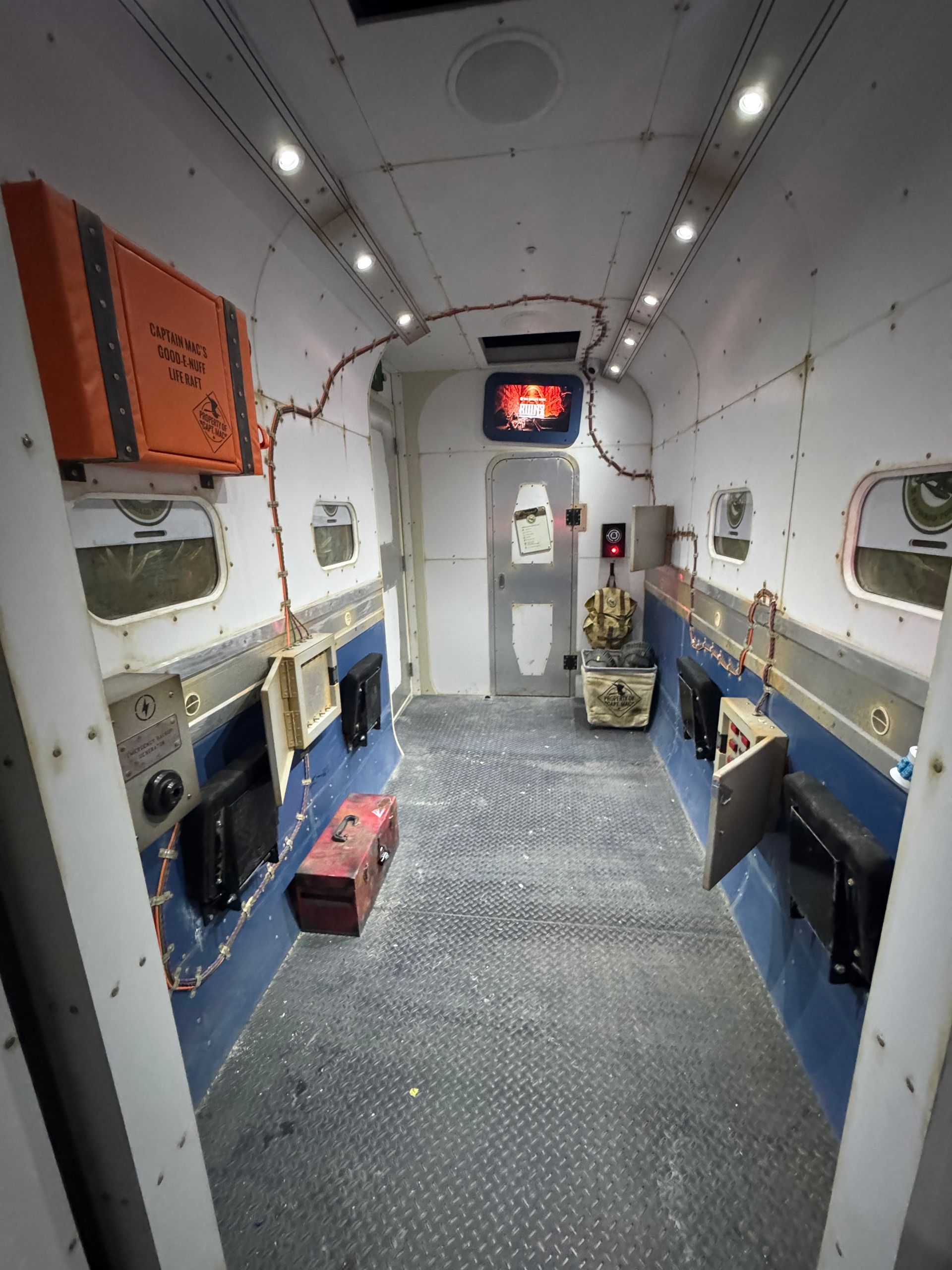 Interior of a narrow, industrial-looking hallway with various equipment, including a red box and screens.