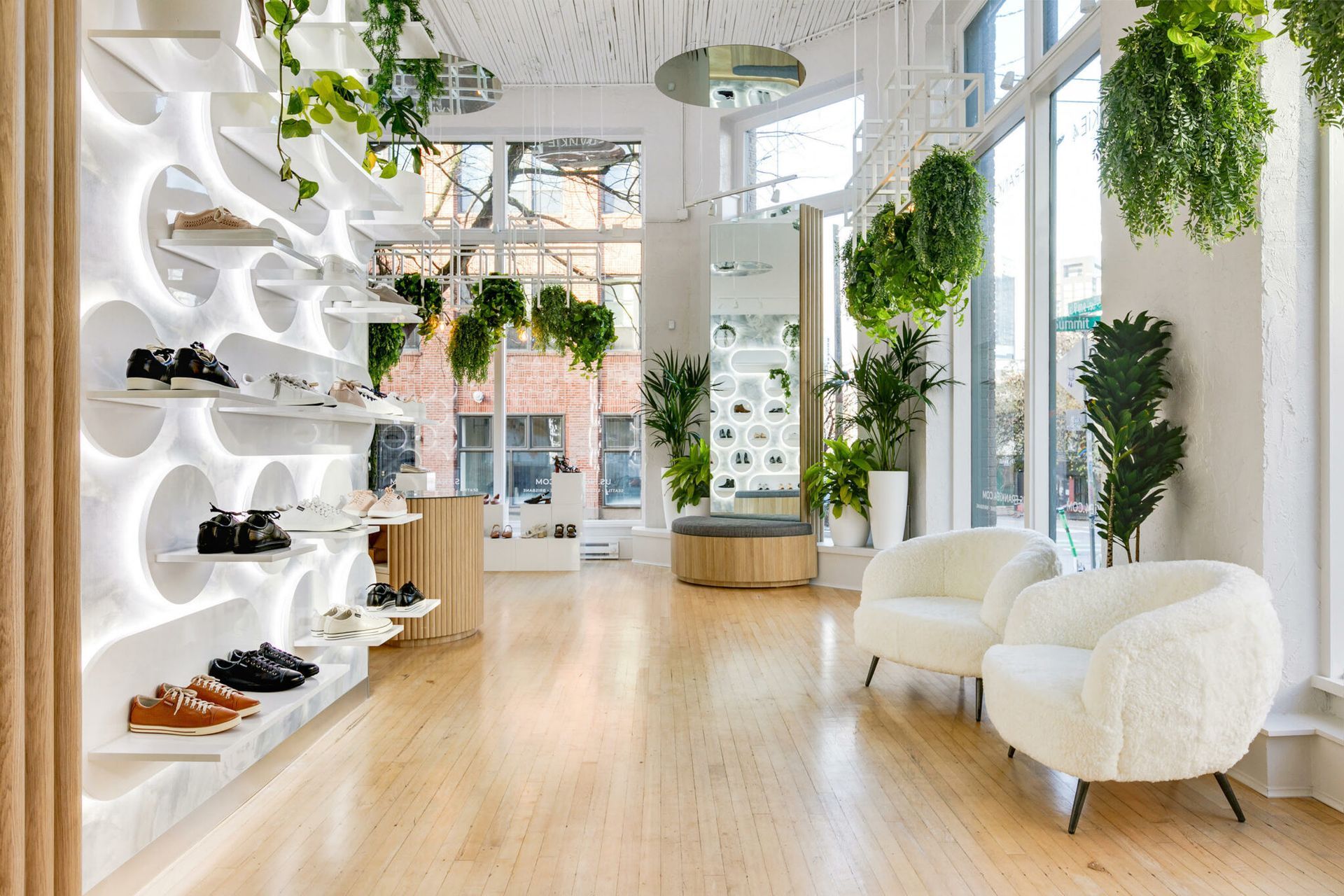 Shoe store interior with wooden floors, white shelving, and hanging plants; two white armchairs sit near windows.