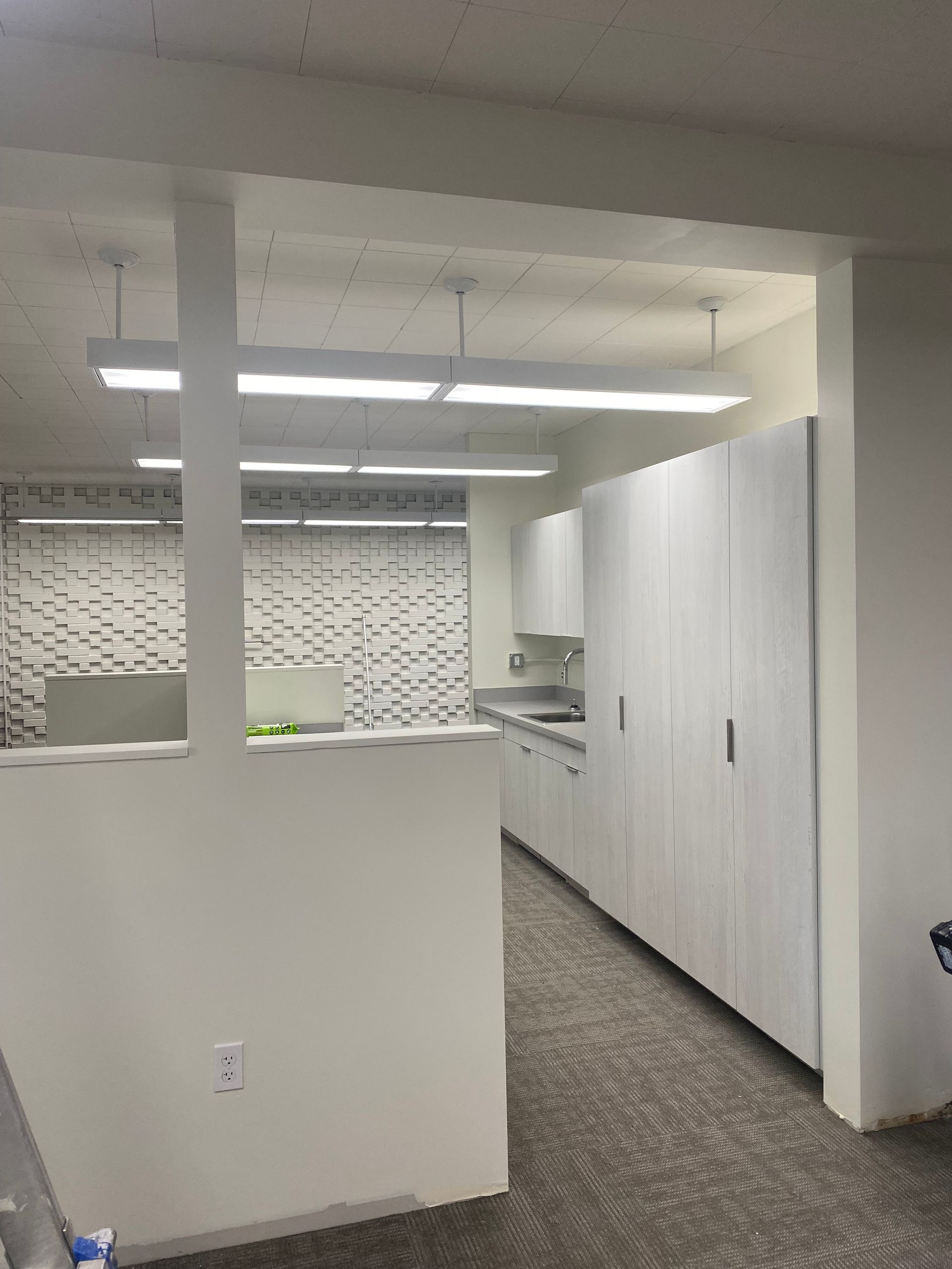 White office space with cabinets, long fluorescent lights, and textured wall visible through an opening.