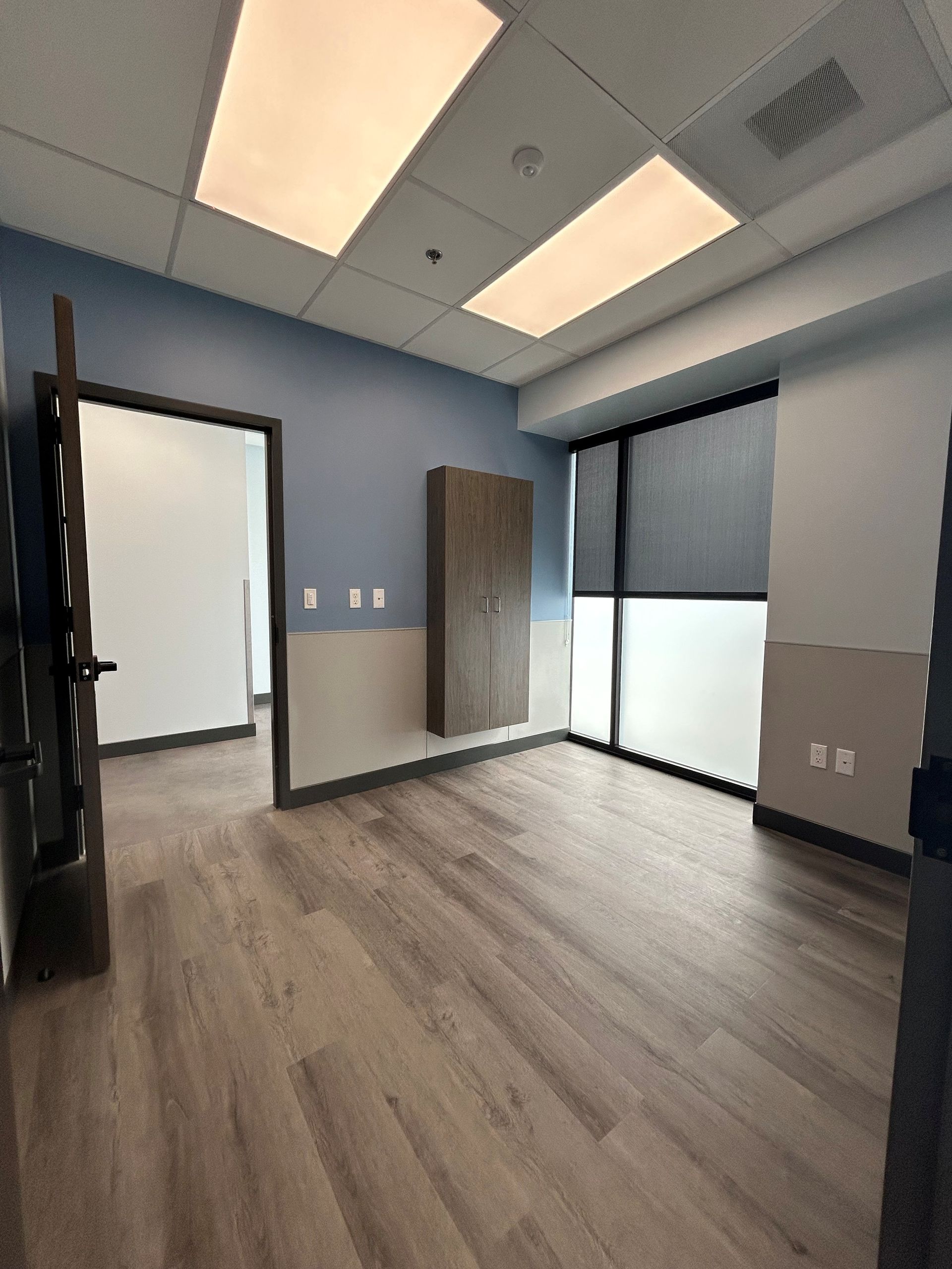 Empty office with wood-look floors, blue and neutral walls, and large windows with blinds.