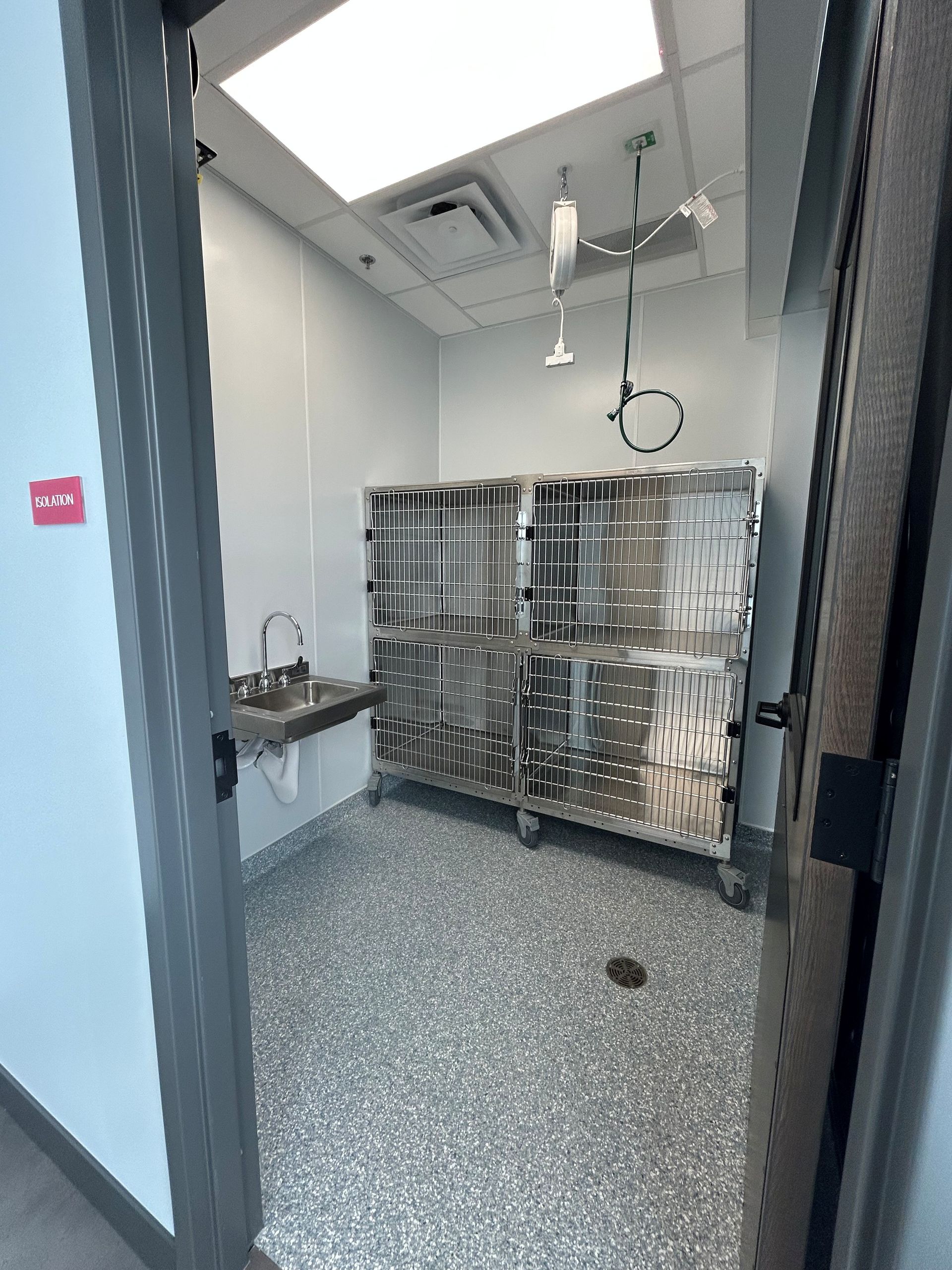 A stainless steel cage on wheels in a room with a sink and overhead medical equipment.