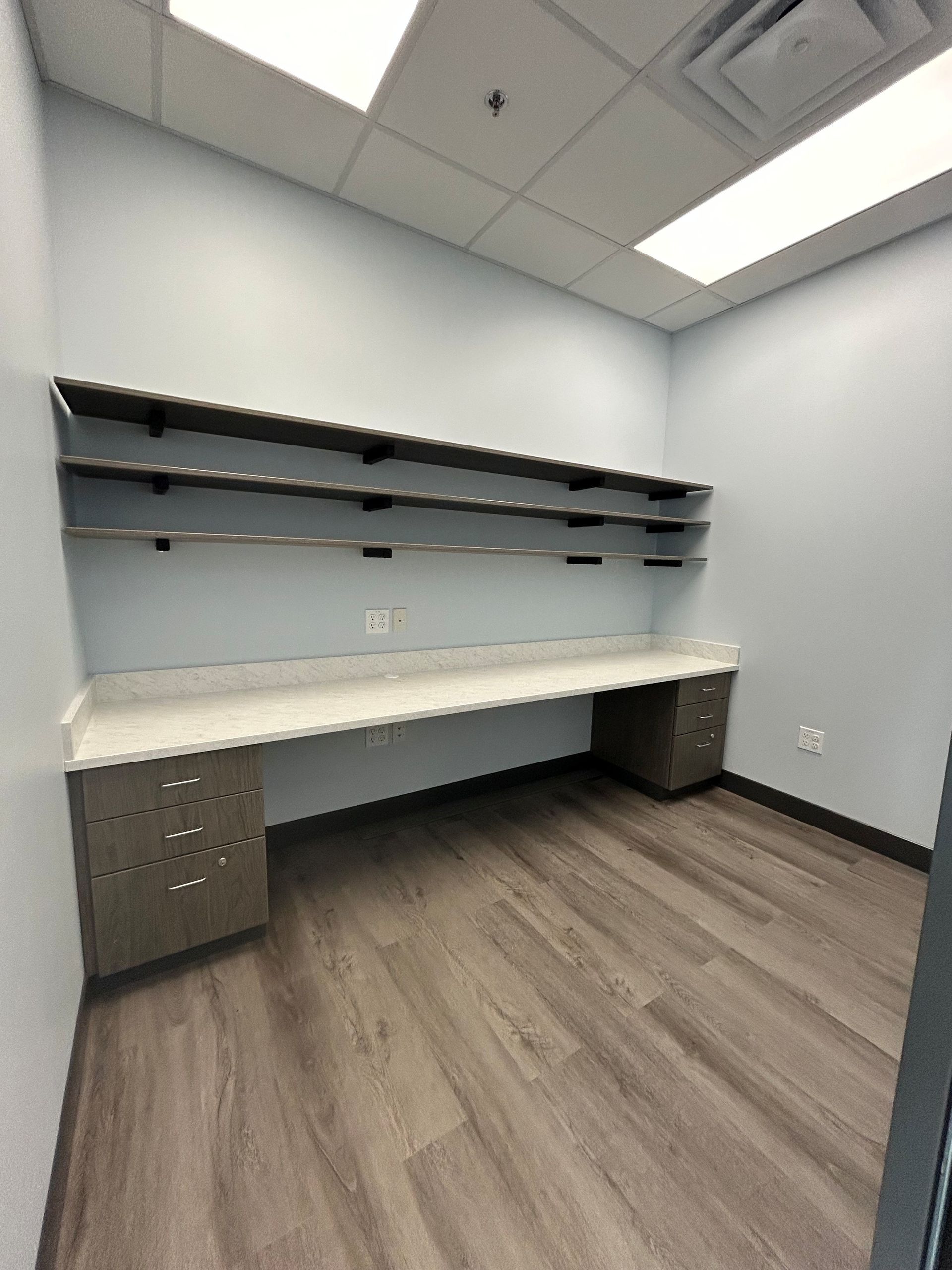 Empty office with built-in desk, shelving, and wood-look flooring. Light blue walls and white ceiling with recessed lighting.