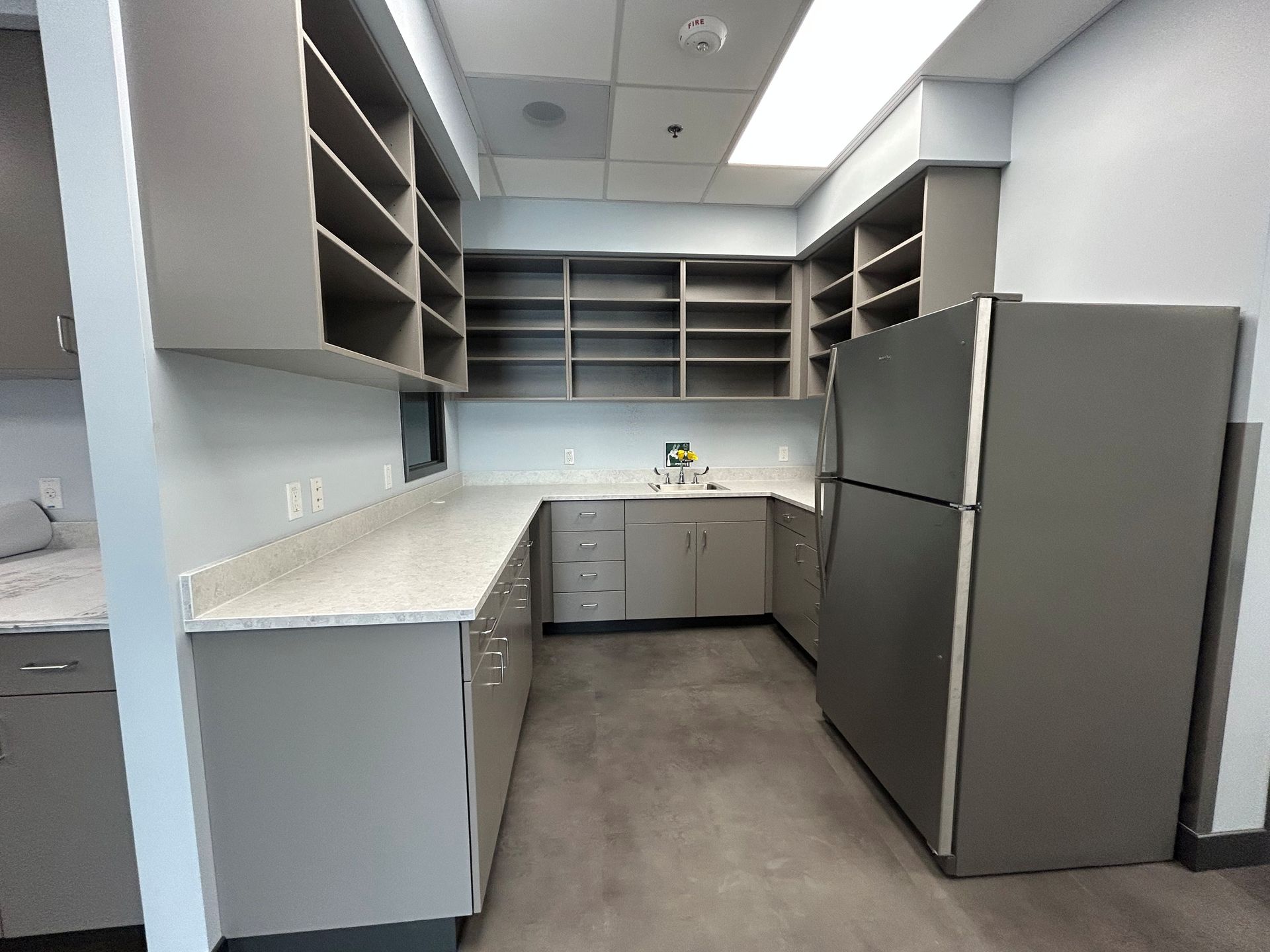 Empty gray kitchen with upper and lower cabinets, counter, refrigerator, and shelving.