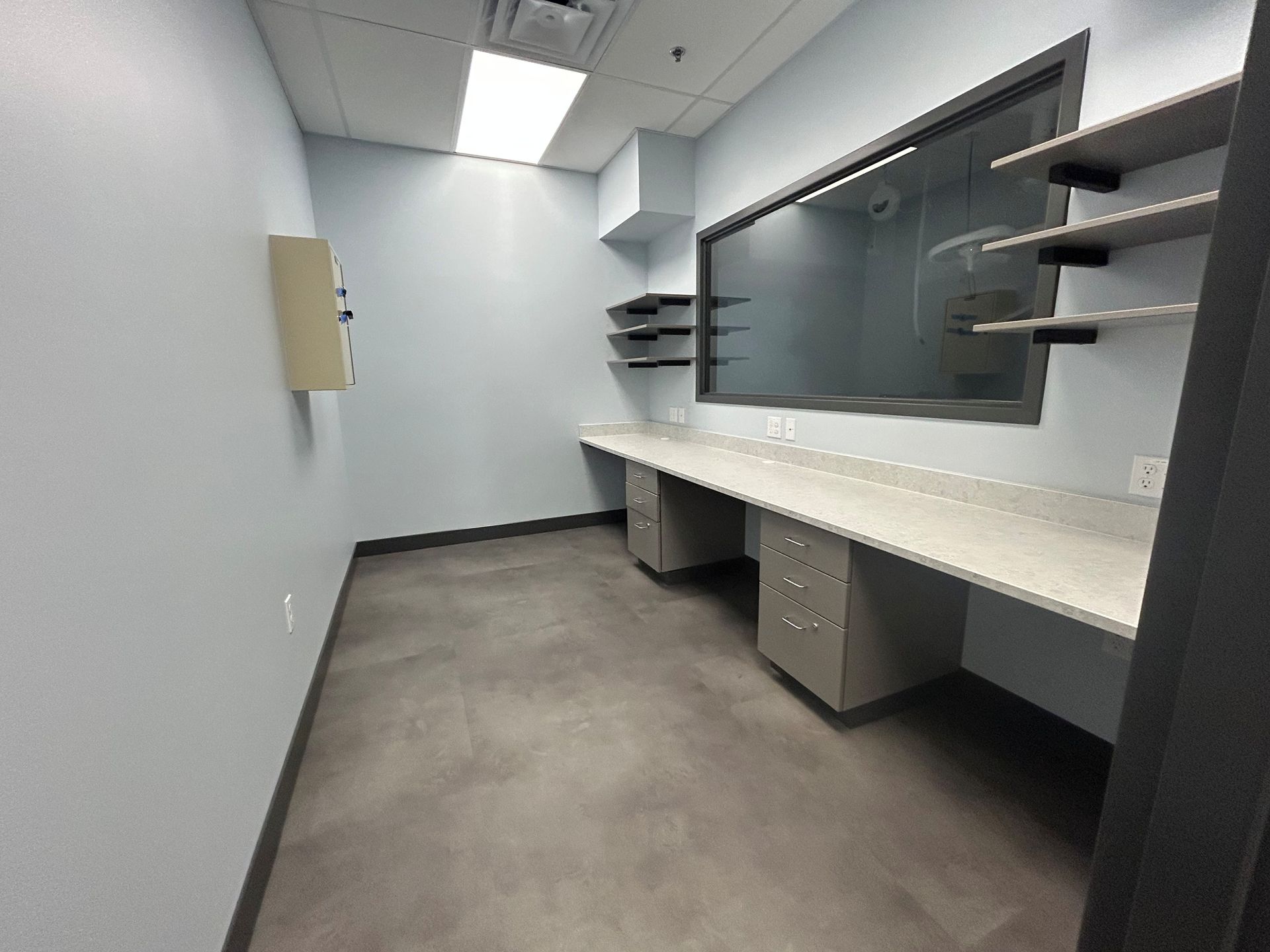 Empty room with long desk, window, and shelves; light blue walls, gray floor, and a closed door.