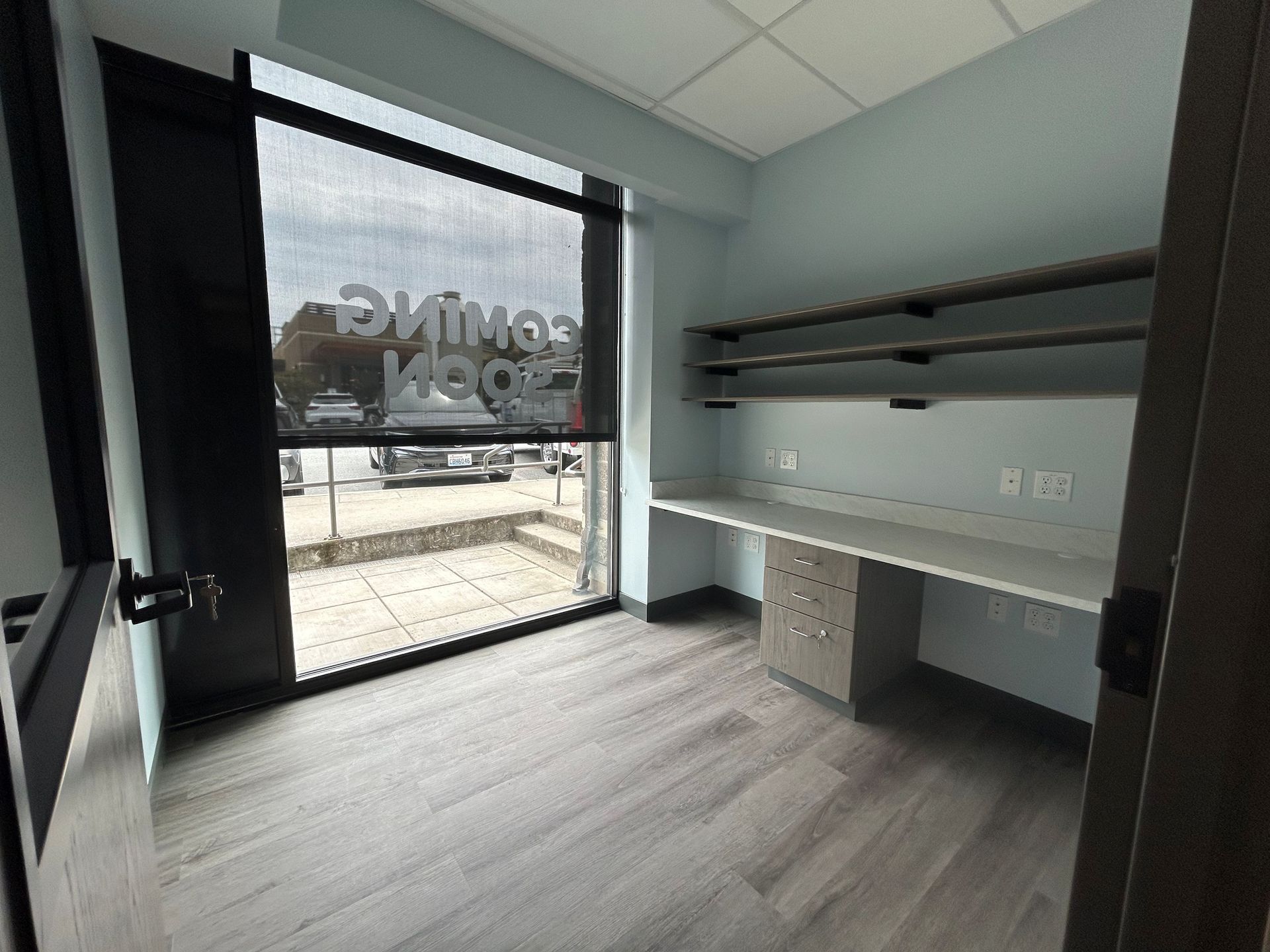 Empty office with a large window, desk, shelves, and gray flooring.