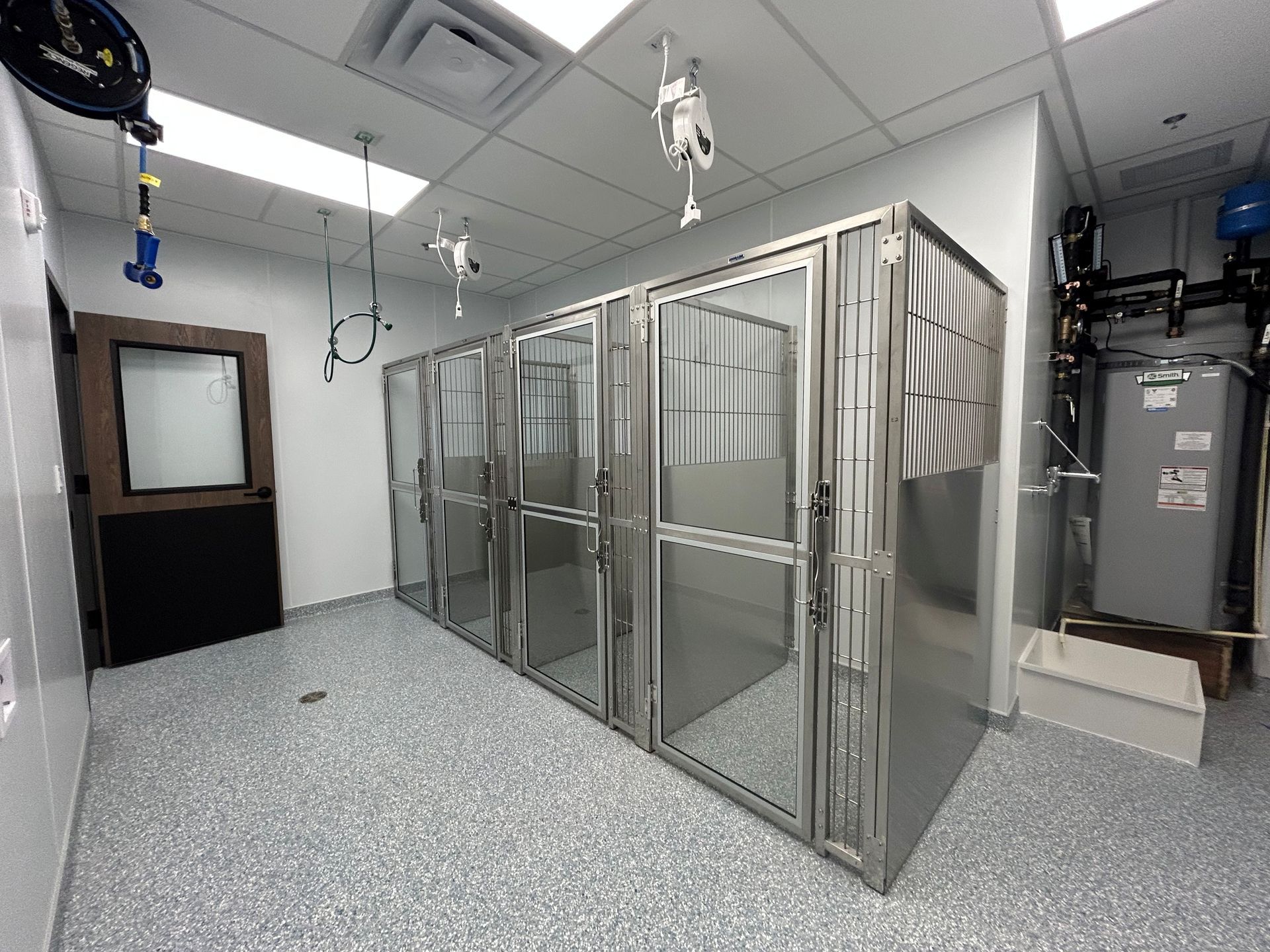 Veterinary clinic with several stainless steel dog kennels and a door. The floor is speckled gray.