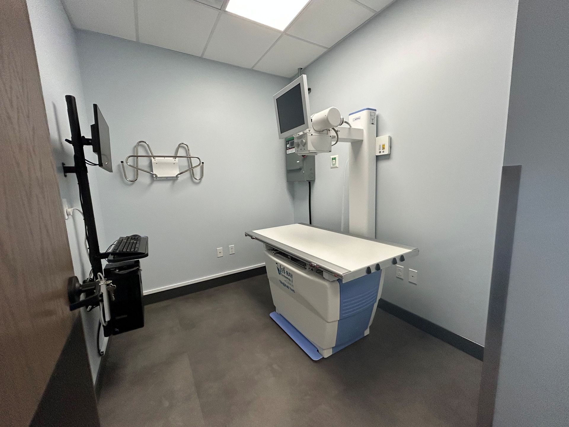 Veterinary radiology room with X-ray machine, white examination table, and monitors. Gray walls and dark flooring.